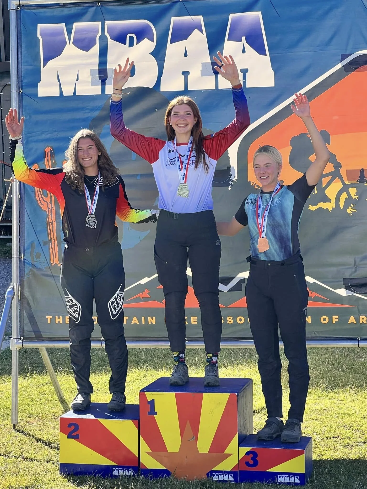 Three female mountain bike racers standing on a winners' podium, holding medals and celebrating. The woman in the center is on the first-place position, wearing a medal and raising her hands. The woman on the left is on the second-place position, and