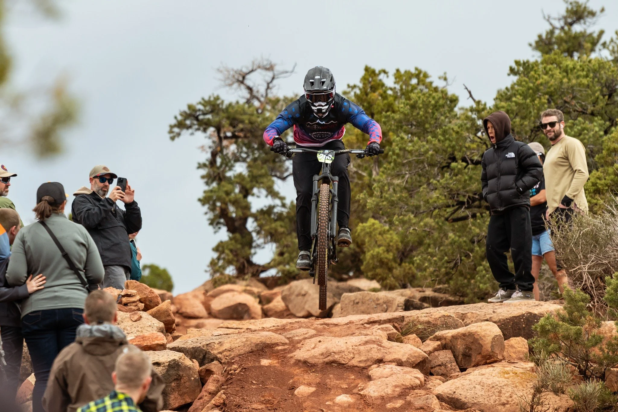 A mountain biker wearing gear and helmet jumping over rocks on a trail, with spectators watching and taking photos