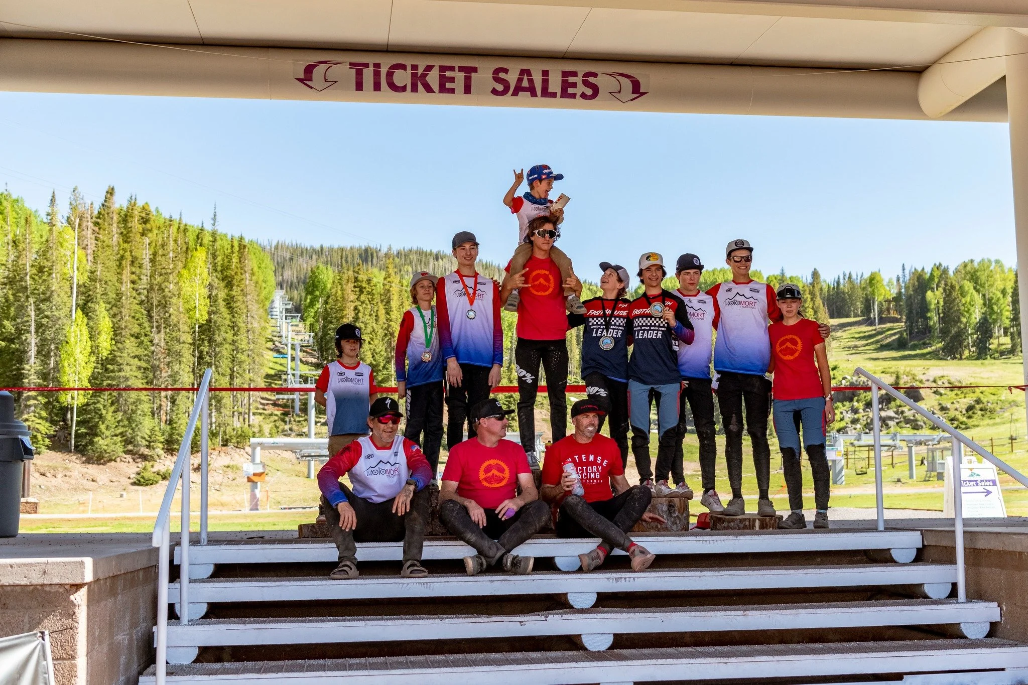 Group of people, mostly young, celebrating with medals and holding a trophy on a podium at a snowy mountain resort, with a ski lift and pine trees in the background.