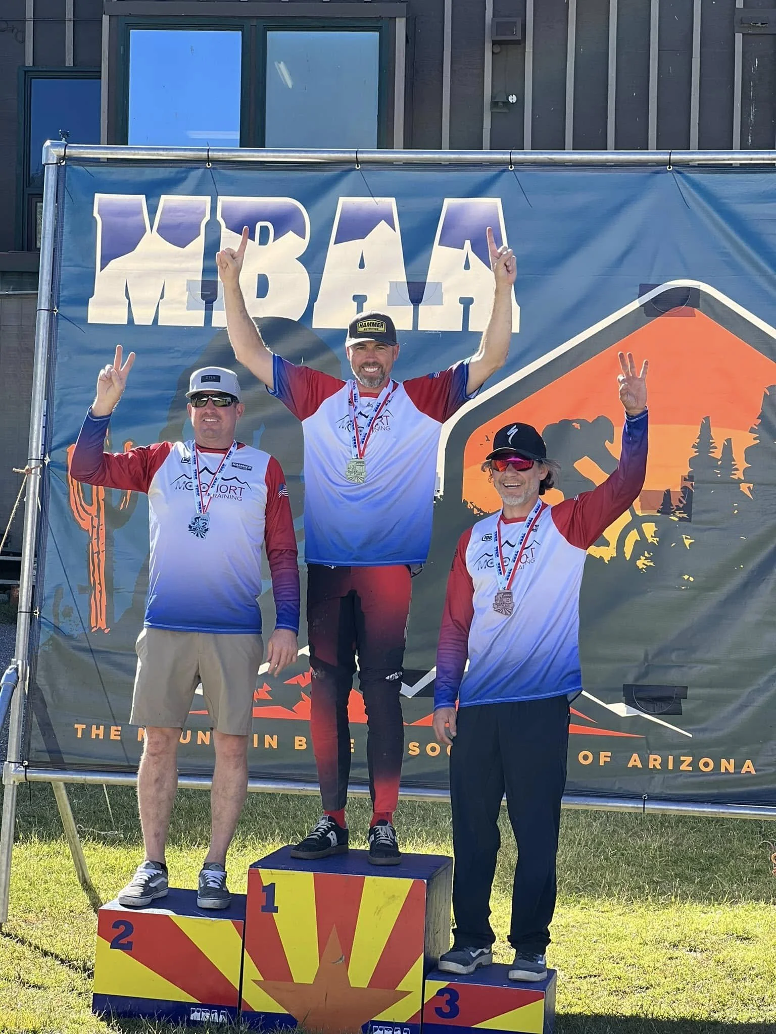 Three men standing on a winners' podium with medals, at the MBRAL event in Arizona. The man in the middle, on the first-place podium, is raising both arms in victory. The other two men are on either side, also holding up peace signs and wearing medal