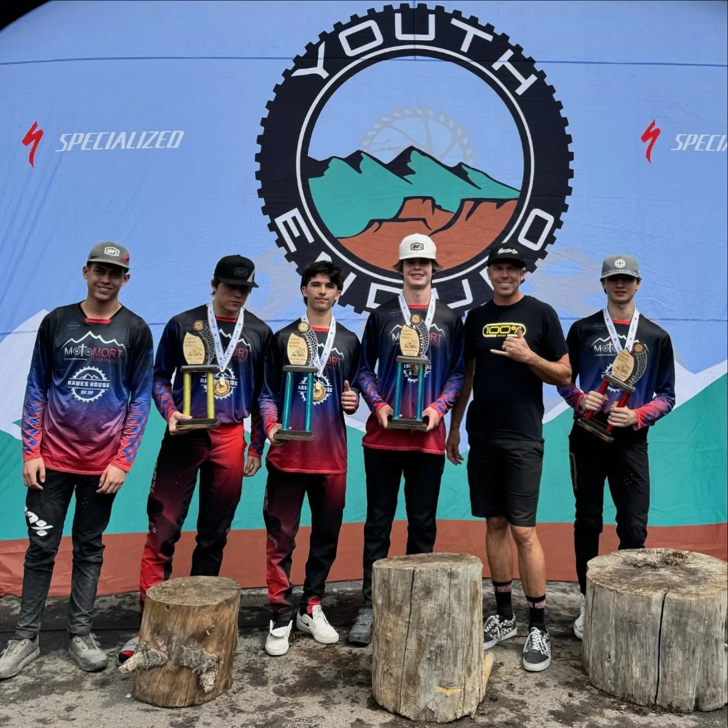 Six young male mountain bikers in matching team jerseys and three adult men standing in front of a large BMX event banner. The boys hold trophies and medals, smiles, and thumbs-up, celebrating a victory. The banner displays a mountain landscape and t
