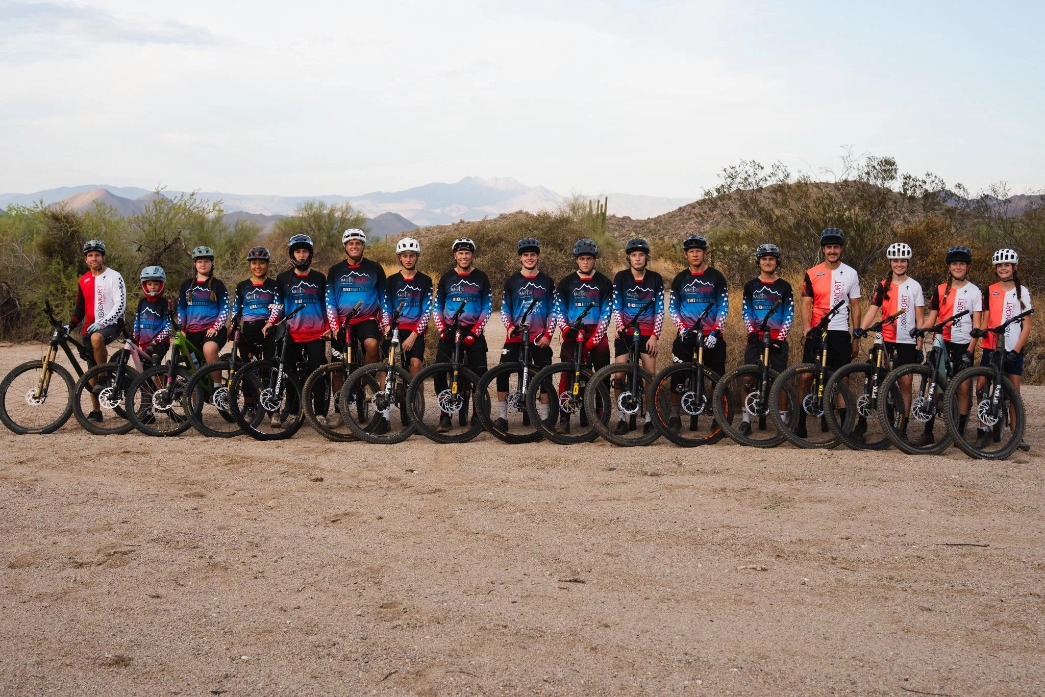 Group of cyclists in matching jerseys and helmets standing with their mountain bikes on a desert trail with mountains and cacti in the background.