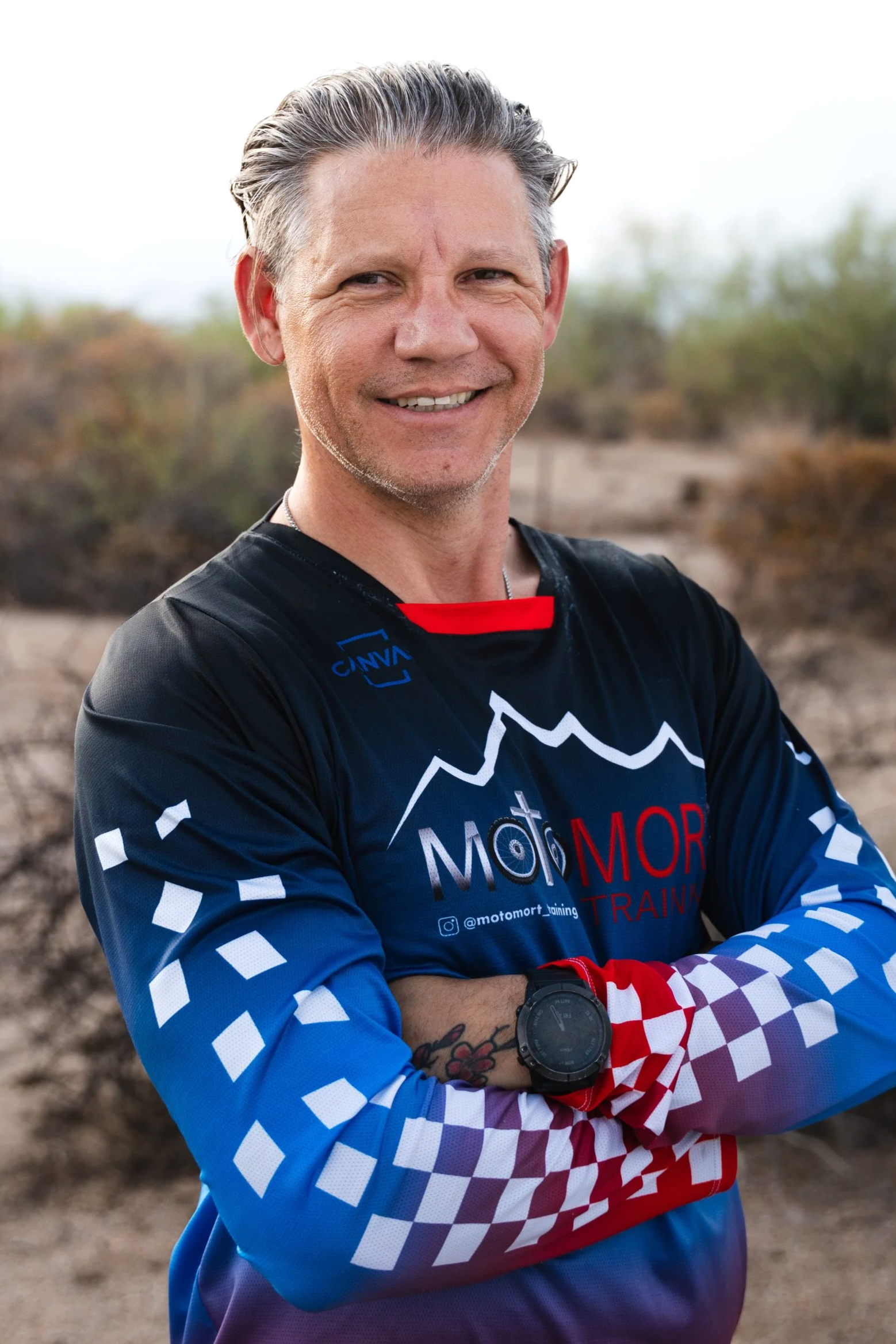 A smiling man wearing a black and blue sports jersey with red and white accents, standing outdoors with a desert landscape in the background.