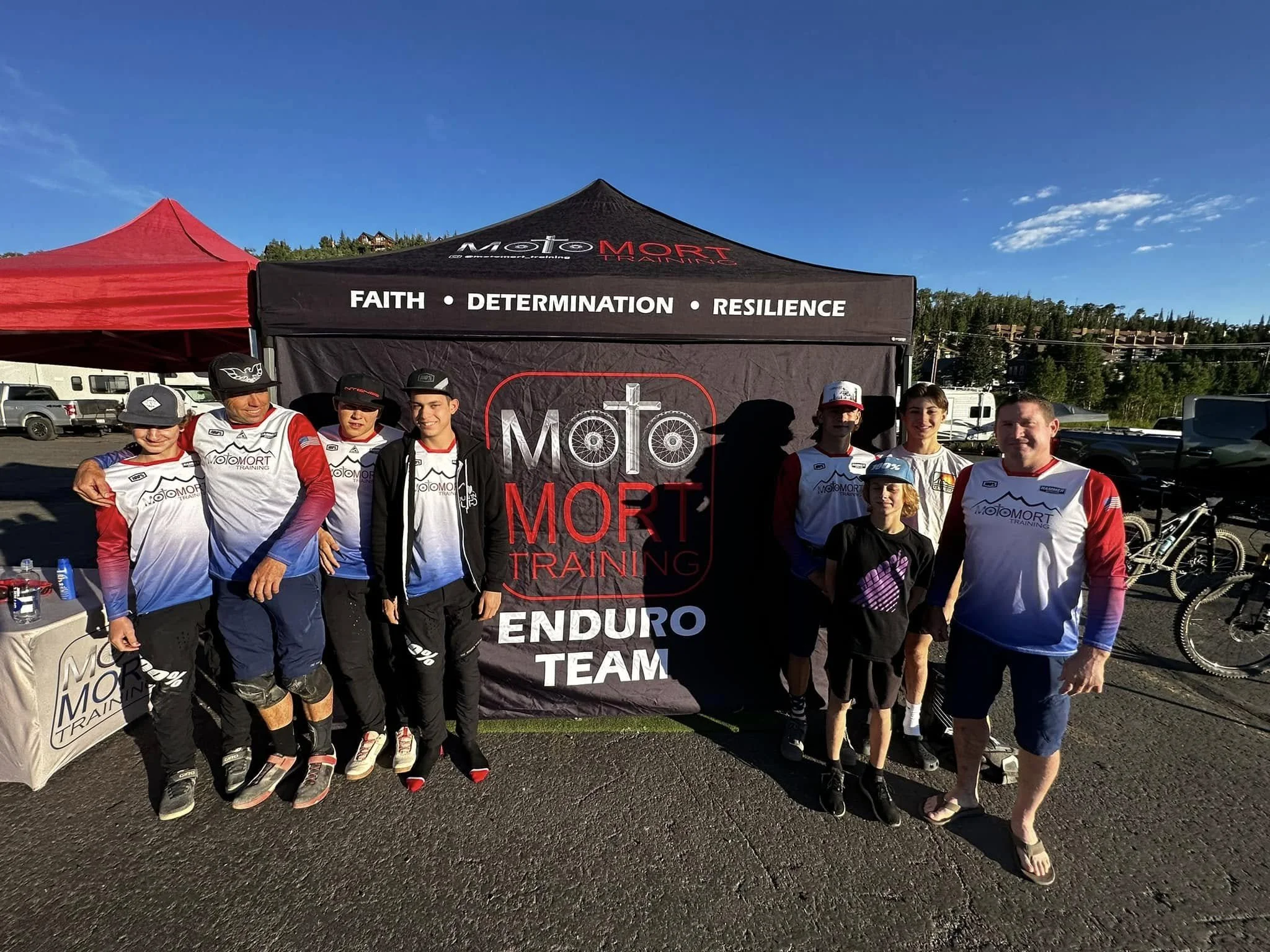 Group of eight people standing in front of a black tent with 'MOTO MORT TRAINING' logo, wearing motocross gear and casual clothing, outdoors on a sunny day.