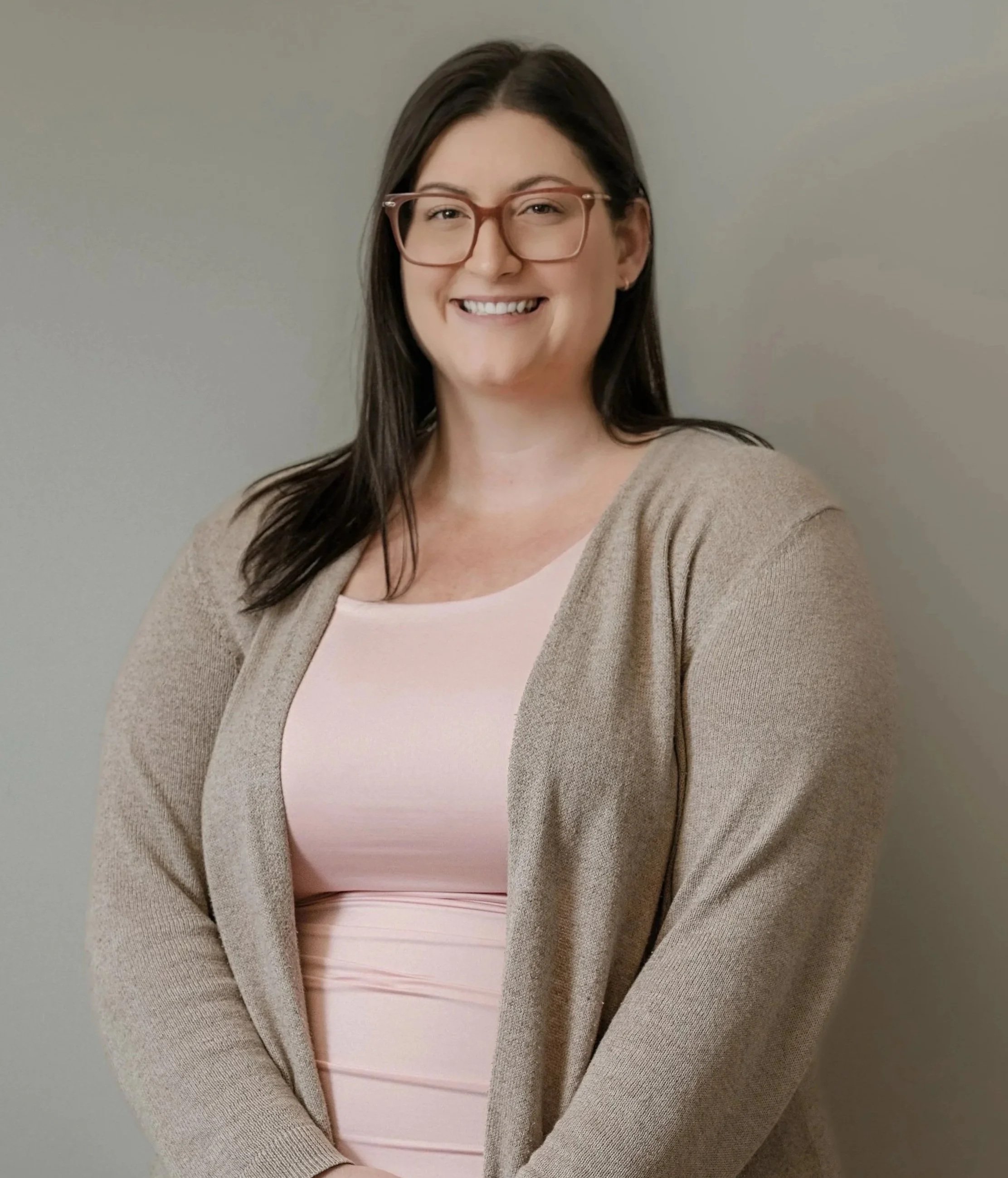 A woman with long brown hair and glasses smiling, wearing a beige cardigan over a light pink top, standing against a neutral background.