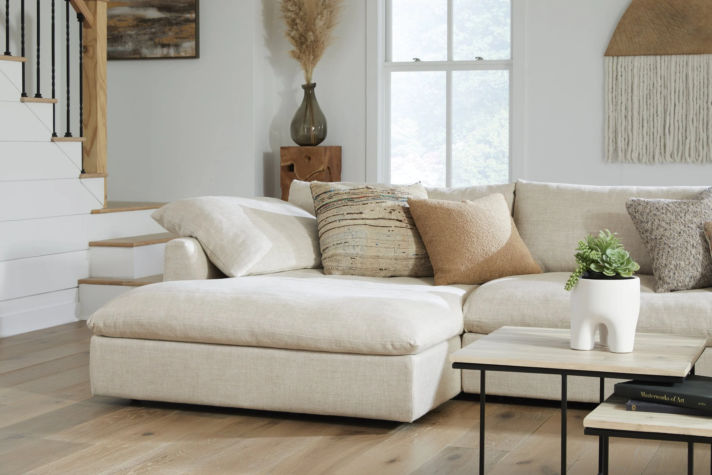 A cozy, modern living room with a large beige sofa decorated with several textured and patterned throw pillows. Next to the sofa is a small wooden coffee table with a white ceramic vase containing a green succulent plant. Behind the sofa, there is a window with white framing allowing natural light in. To the left, part of a staircase with white risers and wooden treads is visible, with black metal balusters. On the wall, linked with the staircase, hangs an abstract painting. To the right of the window, a woven wall hanging and a tall, dark glass vase with dried pampas grass sit atop a sculptural wooden side table. A book titled 'Masterworks of Art' is on a lower shelf of the coffee table.