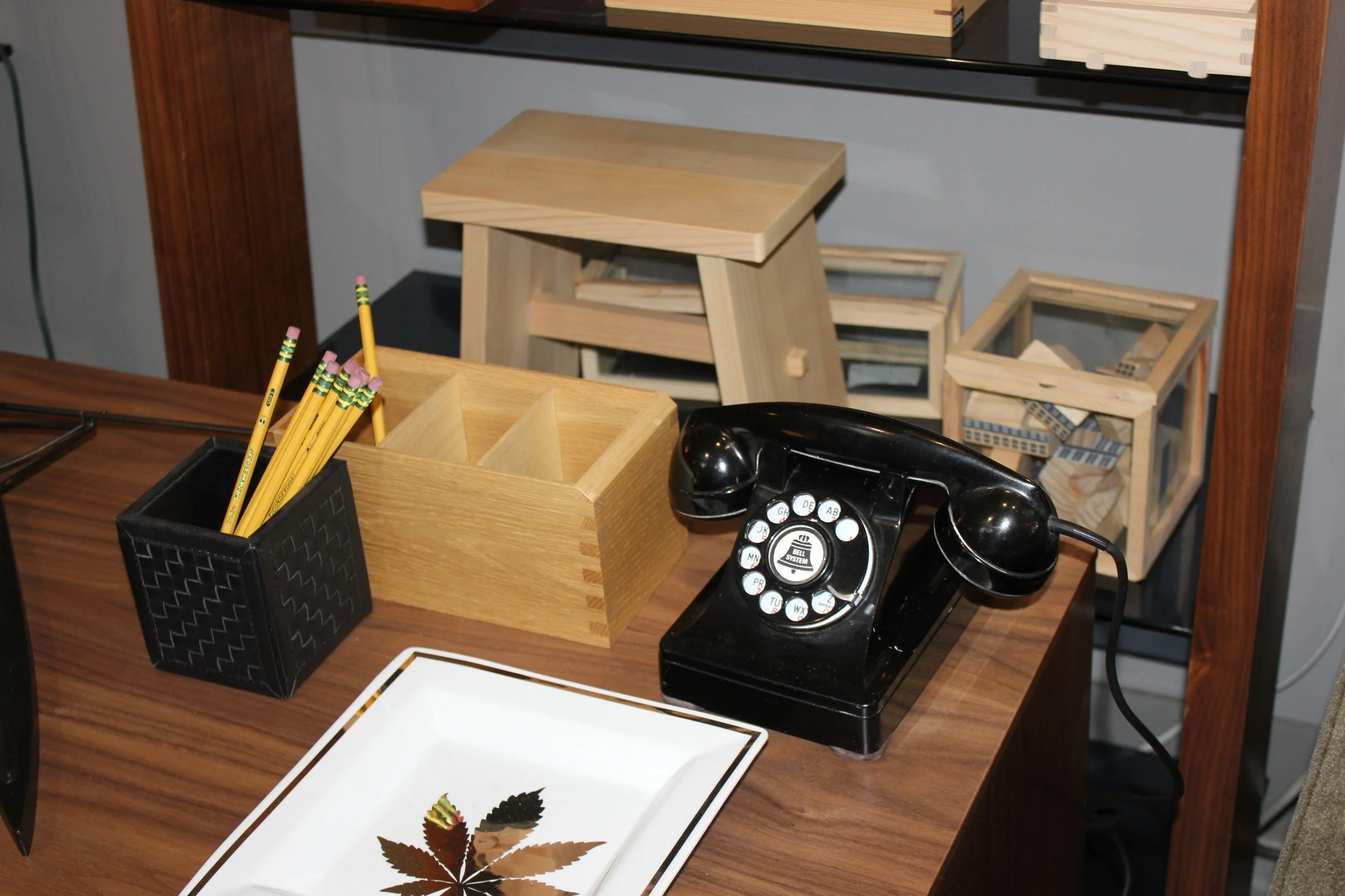 Office desk with a black rotary phone, a white tray with a leaf design, a black pen holder with yellow pencils, a wooden box, and a small wooden frame. In the background, there are extra wooden frames and a shelving unit.