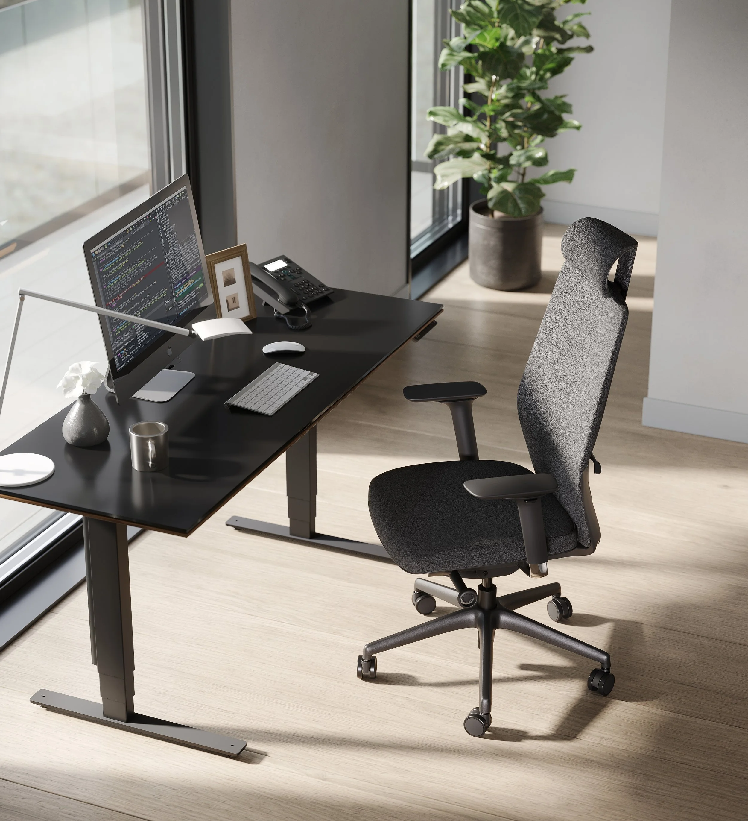 Modern office workspace with a black desk holding a computer monitor displaying code, a keyboard, a mouse, a landline phone, a small framed photo, and a white vase with white flowers. There is an ergonomic office chair with a mesh back, a large potted plant in the background, and a glass window letting in natural light.