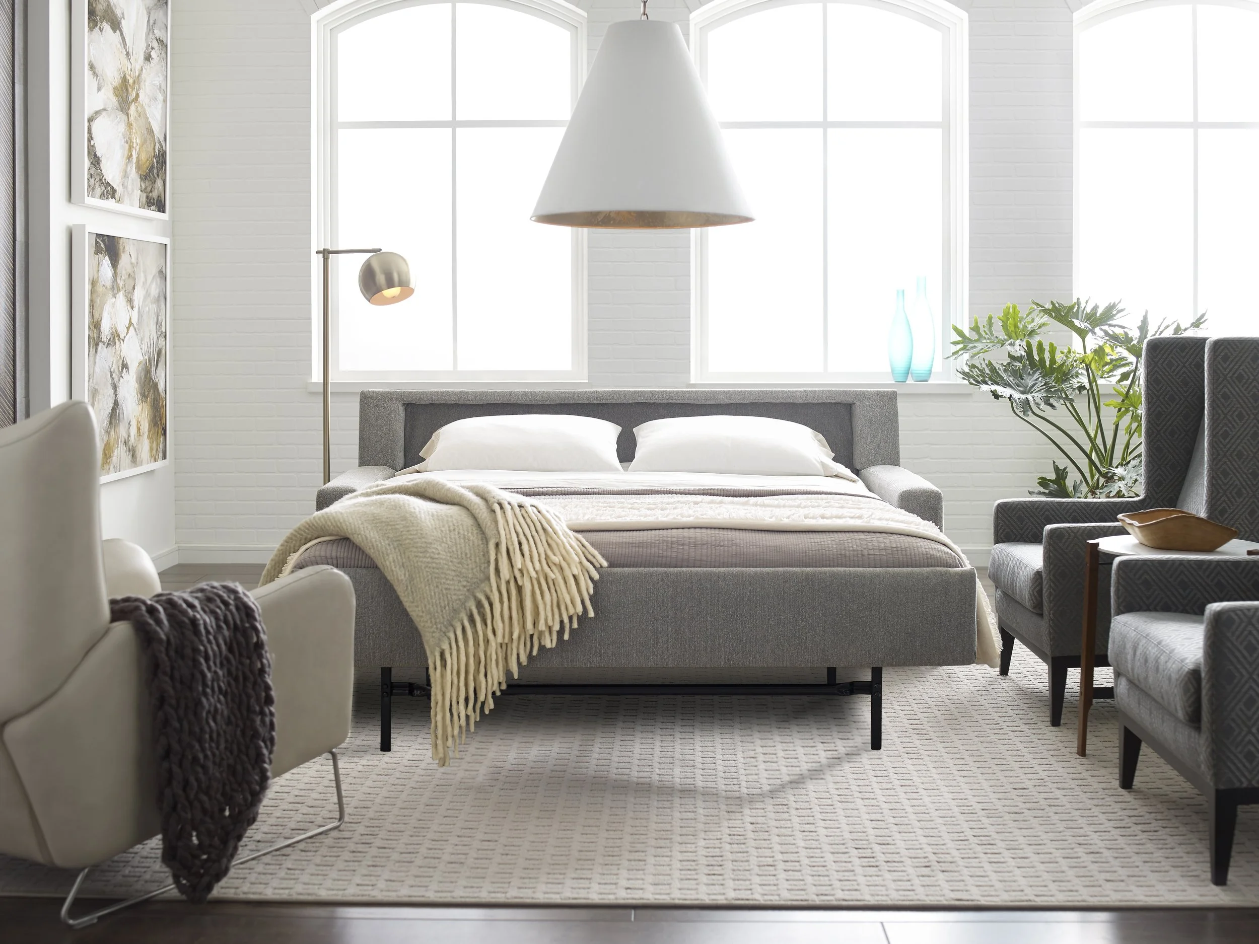 A modern bedroom with white brick walls, large windows, a gray bed, beige and white bedding, a beige armchair with a brown throw blanket, a small side table with a wooden bowl, a gray patterned chair, green plants, two blue vases on the window sill, a large white hanging lamp, and a silver floor lamp.