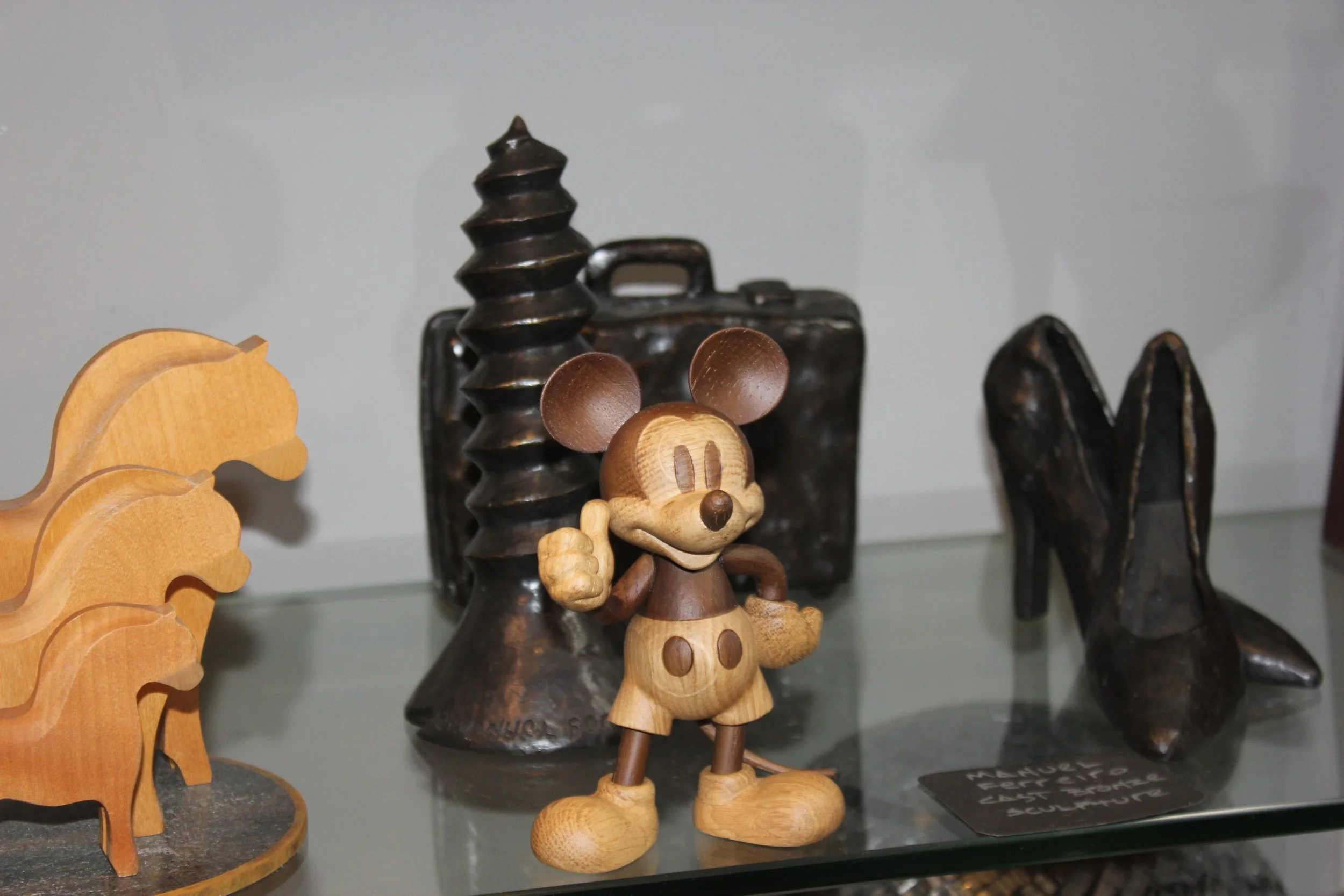 Wooden Mickey Mouse figurine standing in front of a background with metal objects, including a black spiral design, a rectangular metallic box, and some black objects with pointed tips.