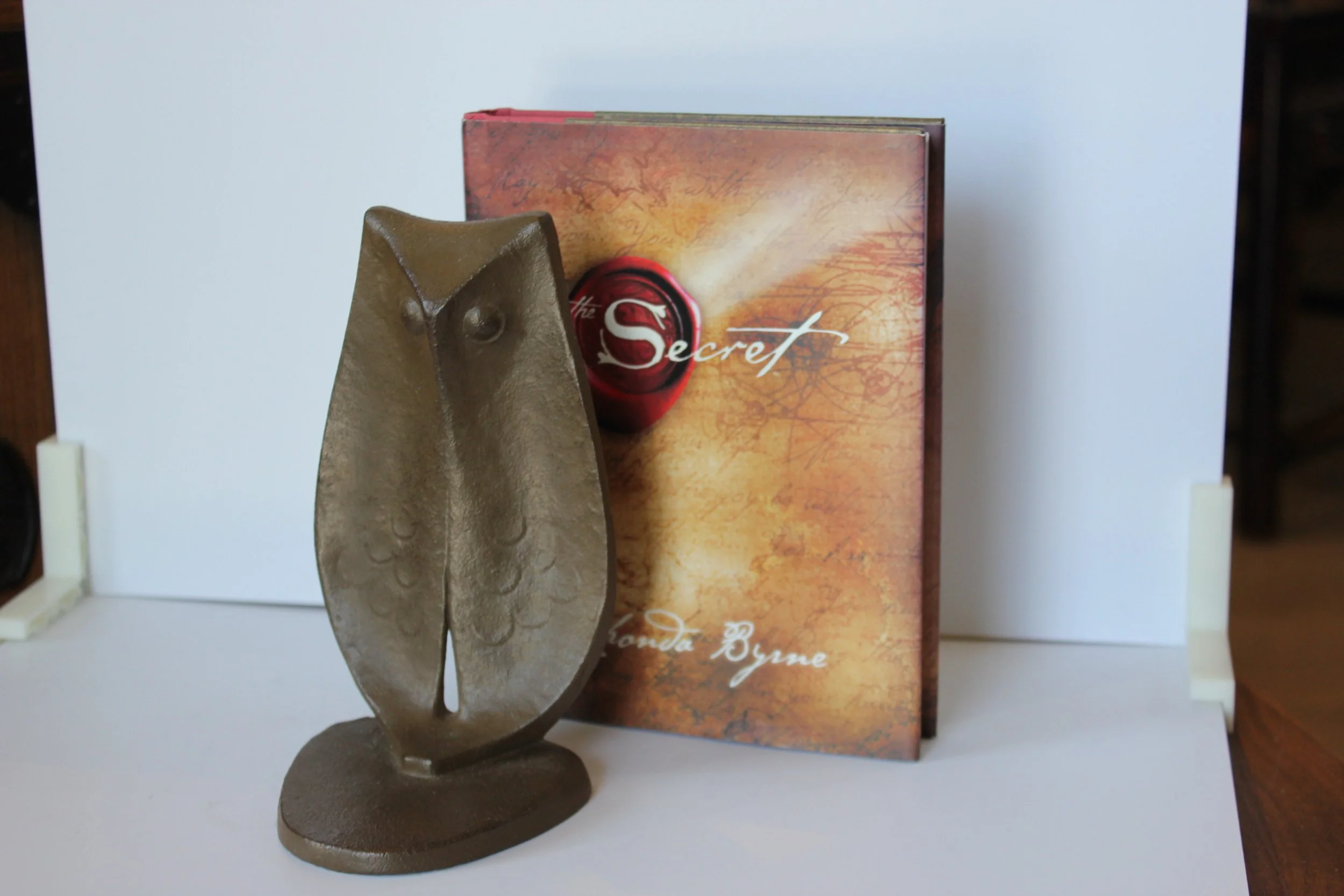 A bronze-colored metal bookmark shaped like a house with a window cutout, placed in front of a book titled 'The Secret' with a red wax seal on the cover, all set against a white background.