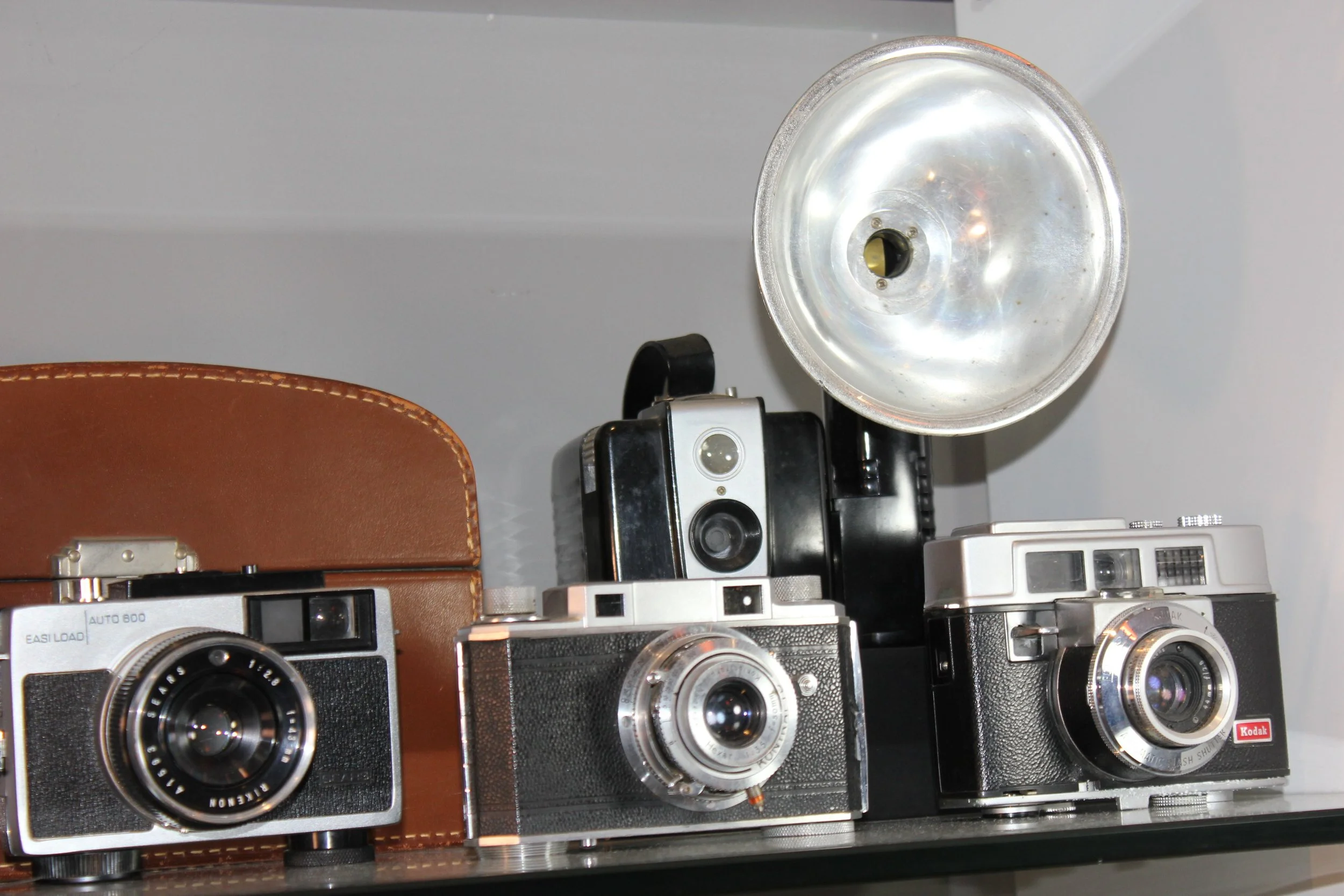 Several vintage cameras placed on a surface, with a silver camera flash and a brown camera case visible among them.