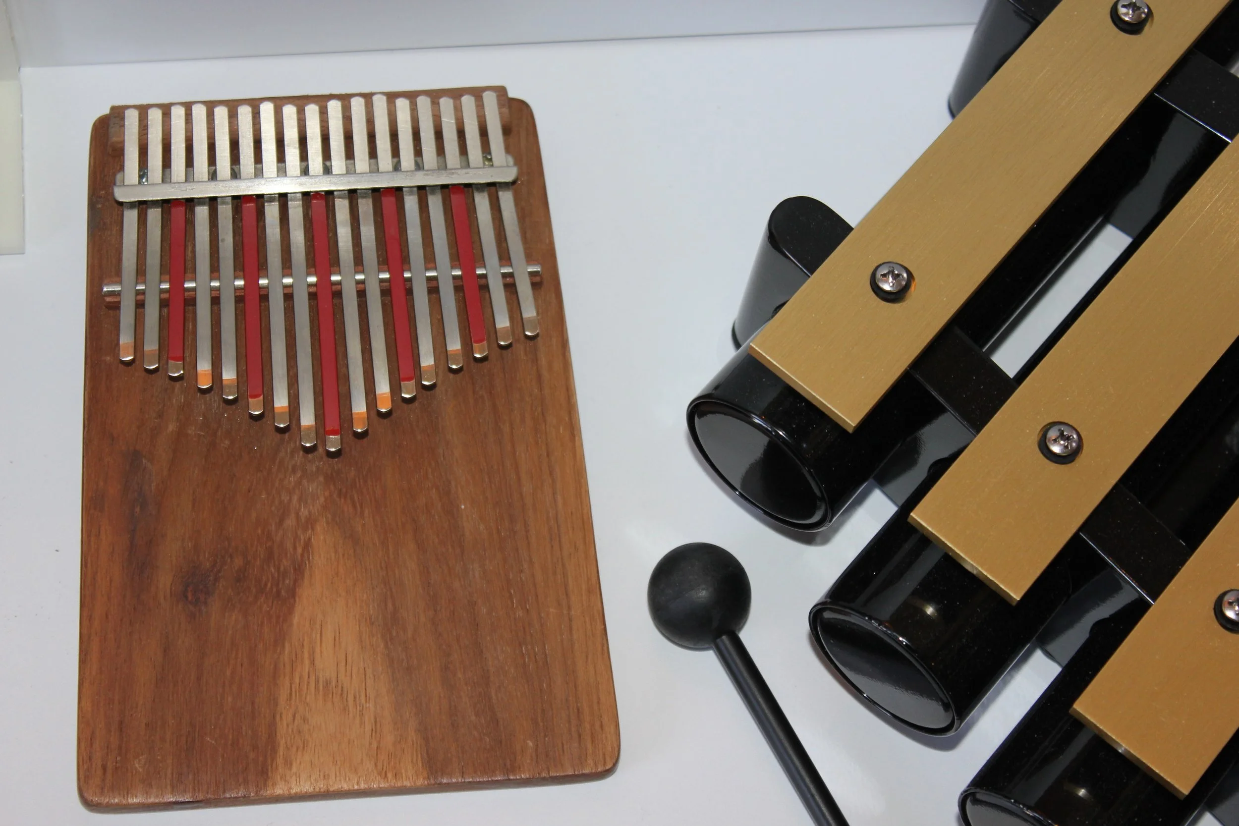 A wooden and metal handheld percussion instrument called a kalimba or thumb piano, with metal tines on a wooden base, is placed on a flat surface with a black and gold percussion instrument and other objects in the background.