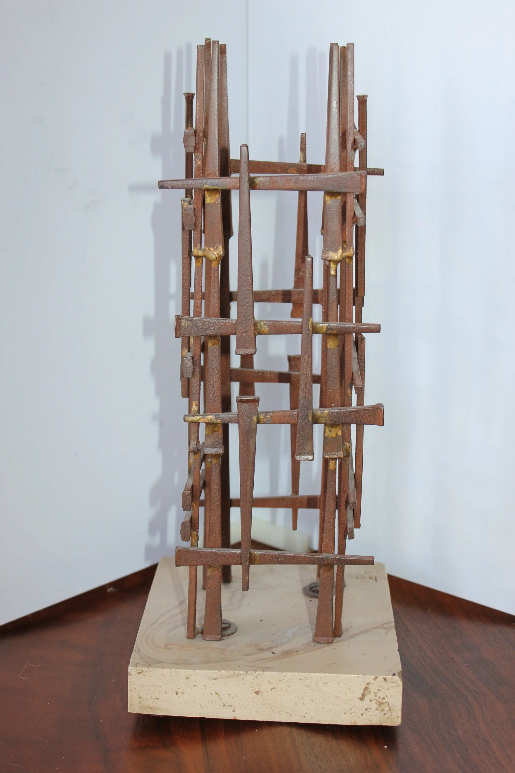 A narrow, tall sculpture made of rusted metal pieces stacked vertically on a white marble base, set against a plain background.