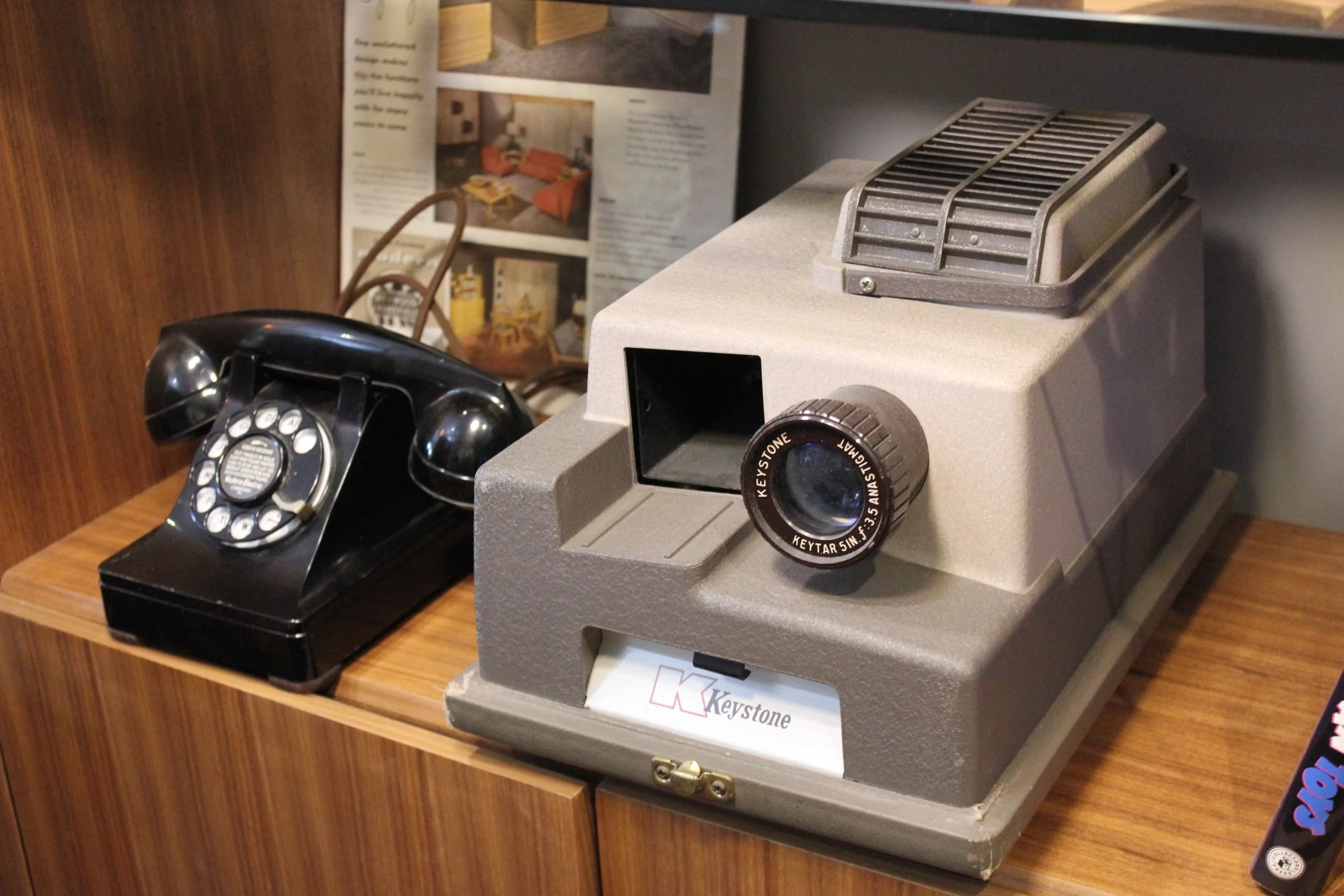 A vintage black rotary telephone sitting on a wooden surface next to a gray vintage microscope labeled 'Keystone' with a large adjustment knob and objective lenses, with a wooden wall and magazine or brochure in the background.