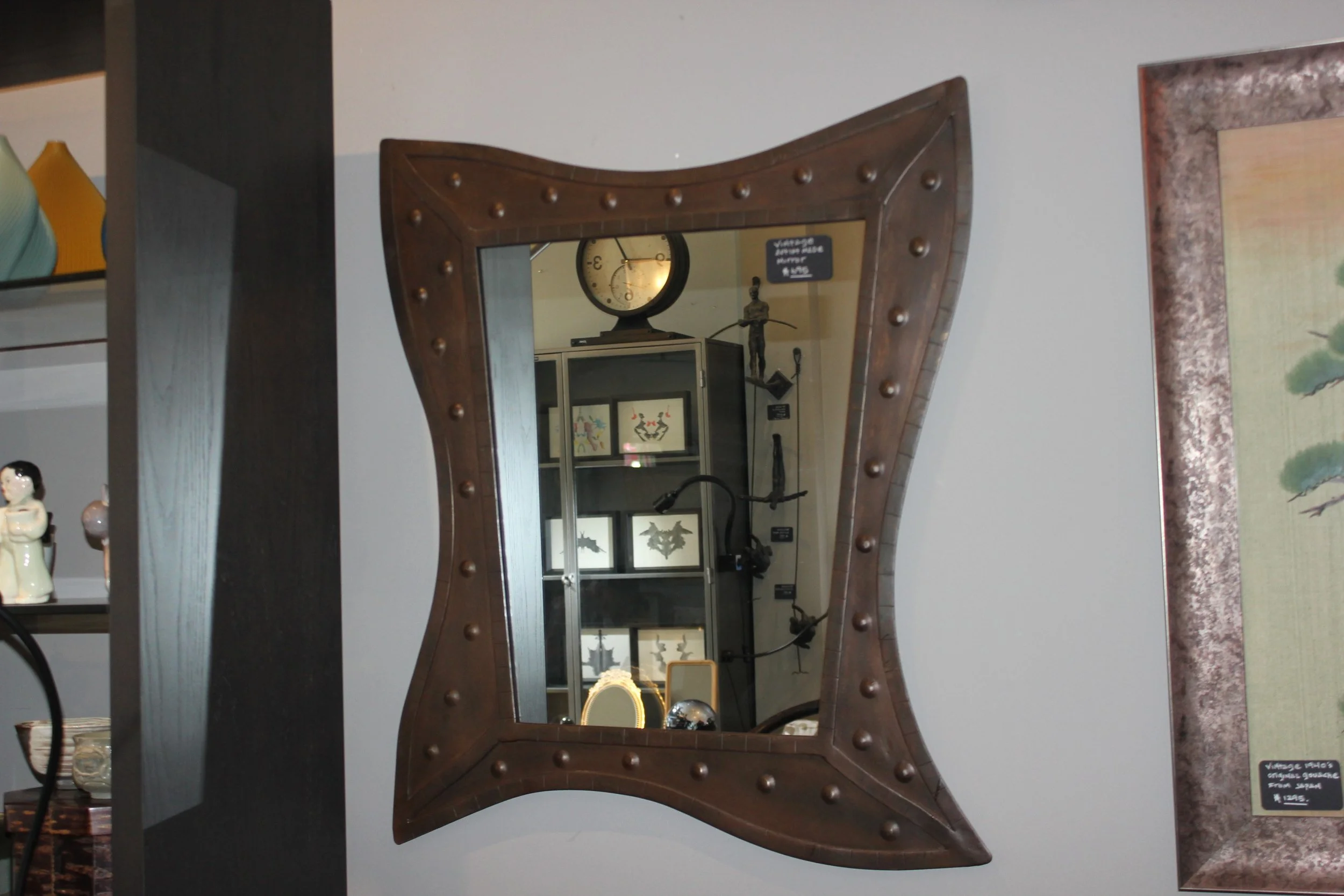 Wooden mirror with round studs, reflecting a clock and decorative items in a room.