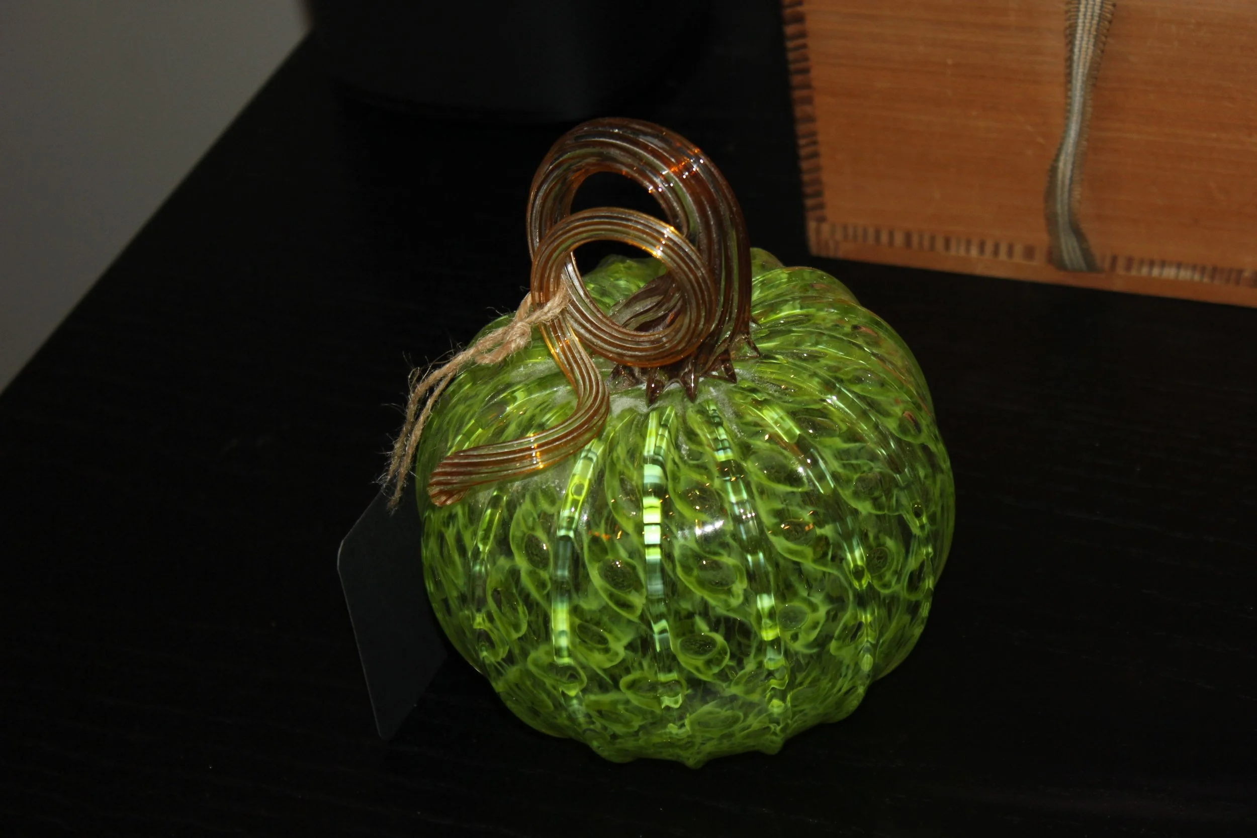 A green glass pumpkin with a spiral glass stem and a textured surface, placed on a dark surface.