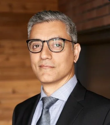 A man with gray hair and glasses wearing a dark suit, light blue shirt, and patterned tie in front of a wooden background.
