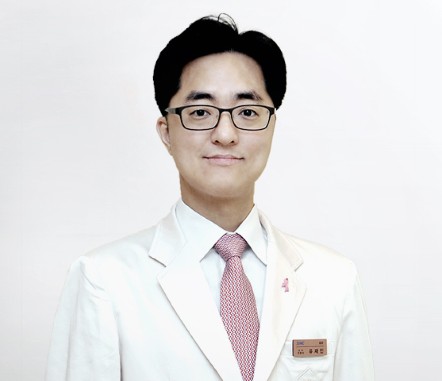 A young man in a white doctor's coat with a pink ribbon pin, glasses, and a patterned pink tie, standing against a plain white background.