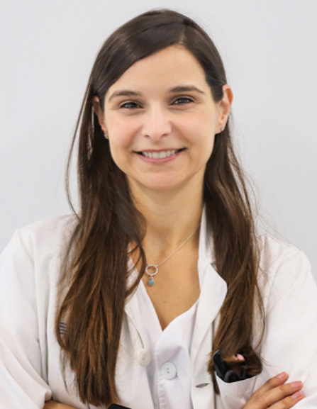 A young woman with long brown hair, dressed in a white coat, smiling with her arms crossed, against a plain light gray background.