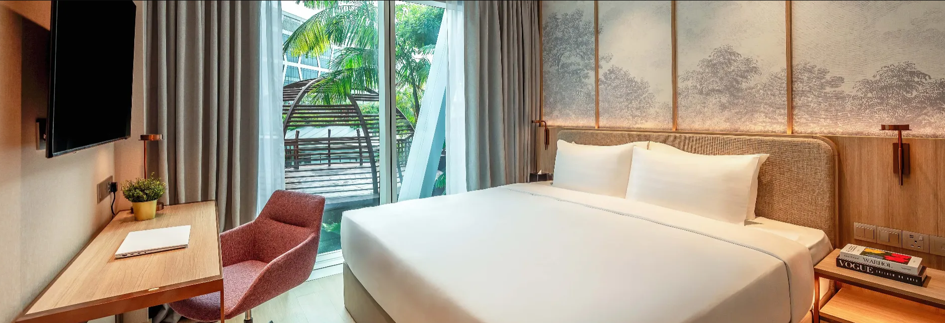 Modern hotel room with a large bed, a bedside table with magazines, a wall-mounted TV, a desk with a pink chair, and a sliding glass door leading to an outdoor balcony with greenery.