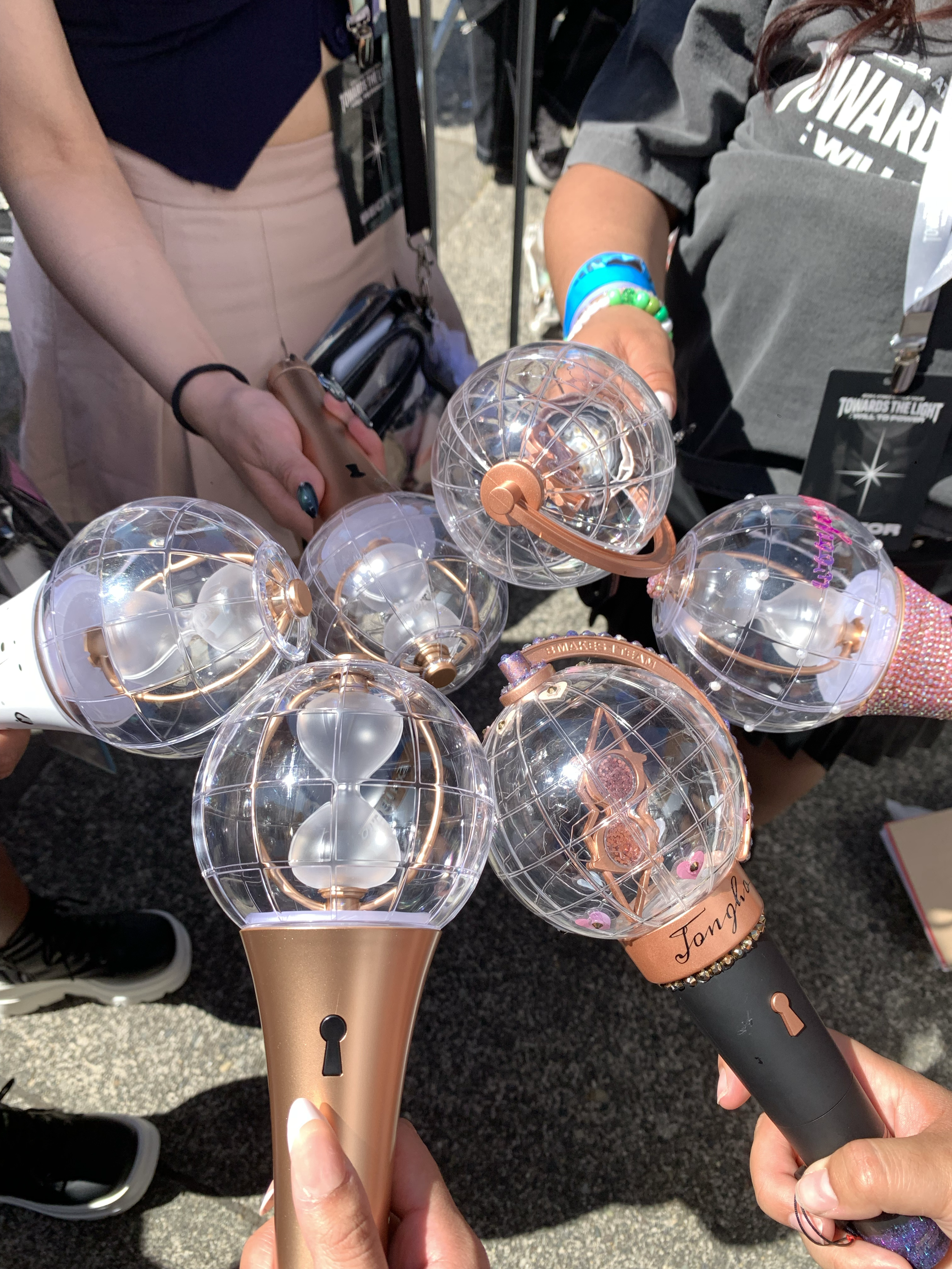Multiple handheld light sticks with a rose gold and black handle, decorated with clear spherical bulbs and rose gold accents, being held by people at an outdoor event.