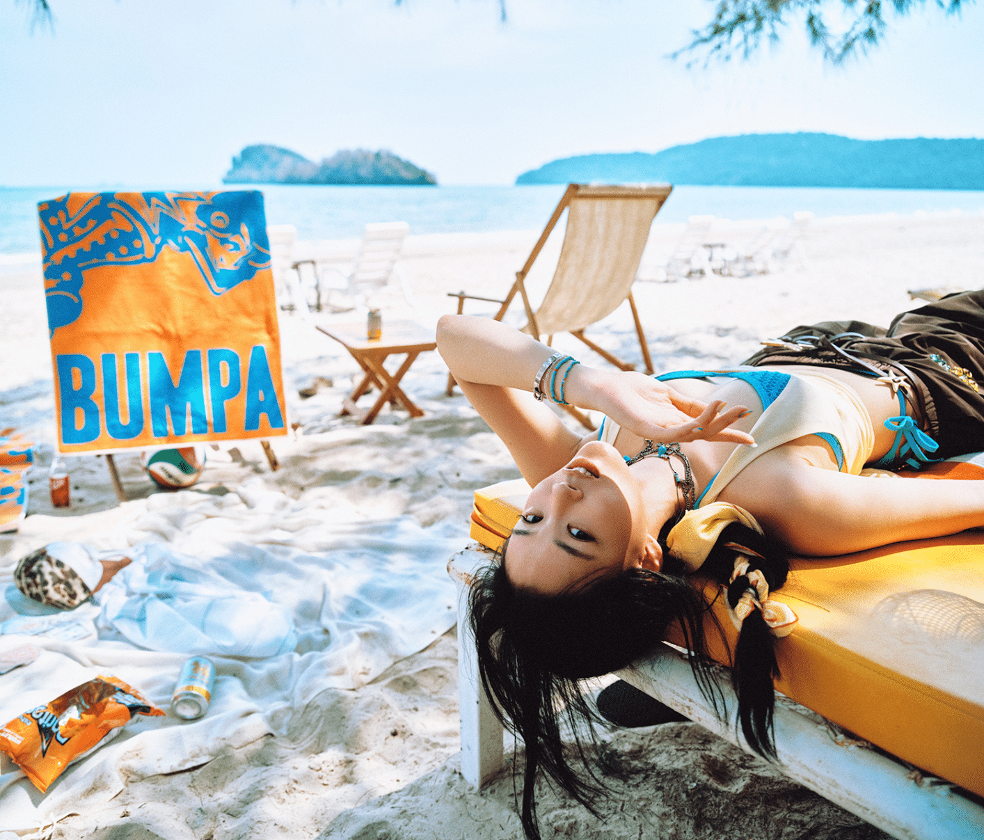 A woman lying on a yellow beach lounge on a sandy beach near the ocean, with islands in the background. She has long dark hair, wears a blue and yellow swimsuit, and has jewelry on her wrist. There are beach chairs, a sign that reads 'BUMP A,' and snacks and drinks scattered on the sand.