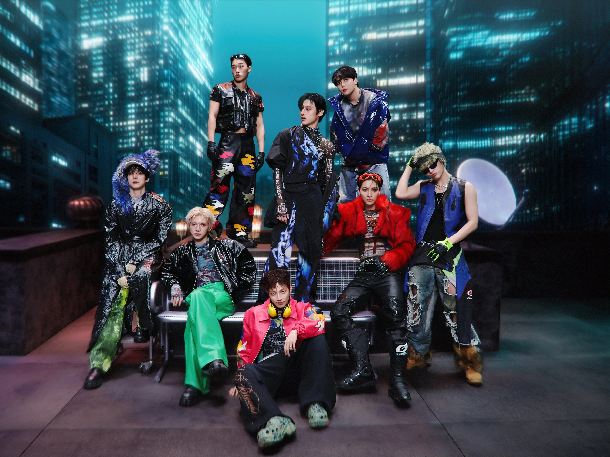 A group of young men in colorful, futuristic, and eclectic outfits with bold patterns, accessories, and hairstyles, pose on a set resembling a cityscape at night with glowing skyscrapers in the background.