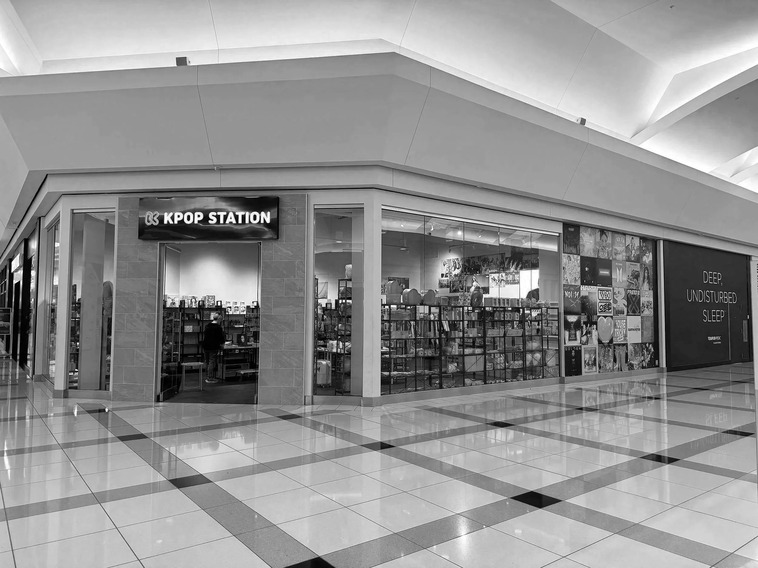 Interior view of a shopping mall, showing a bookstore named 'KPOP STATION' with glass display windows filled with books and posters, tiled floor, and modern architectural ceiling with lighting.