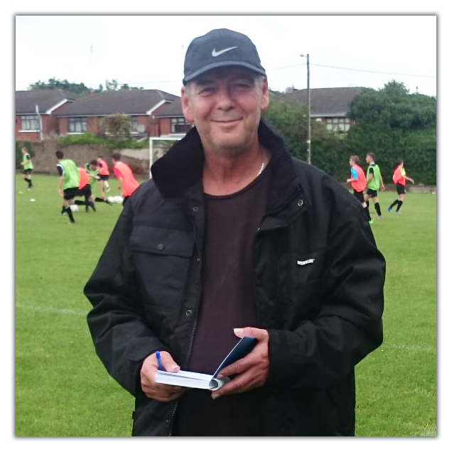 A man wearing a black Nike cap and jacket, holding a notebook, standing on a soccer field with children playing soccer in the background.