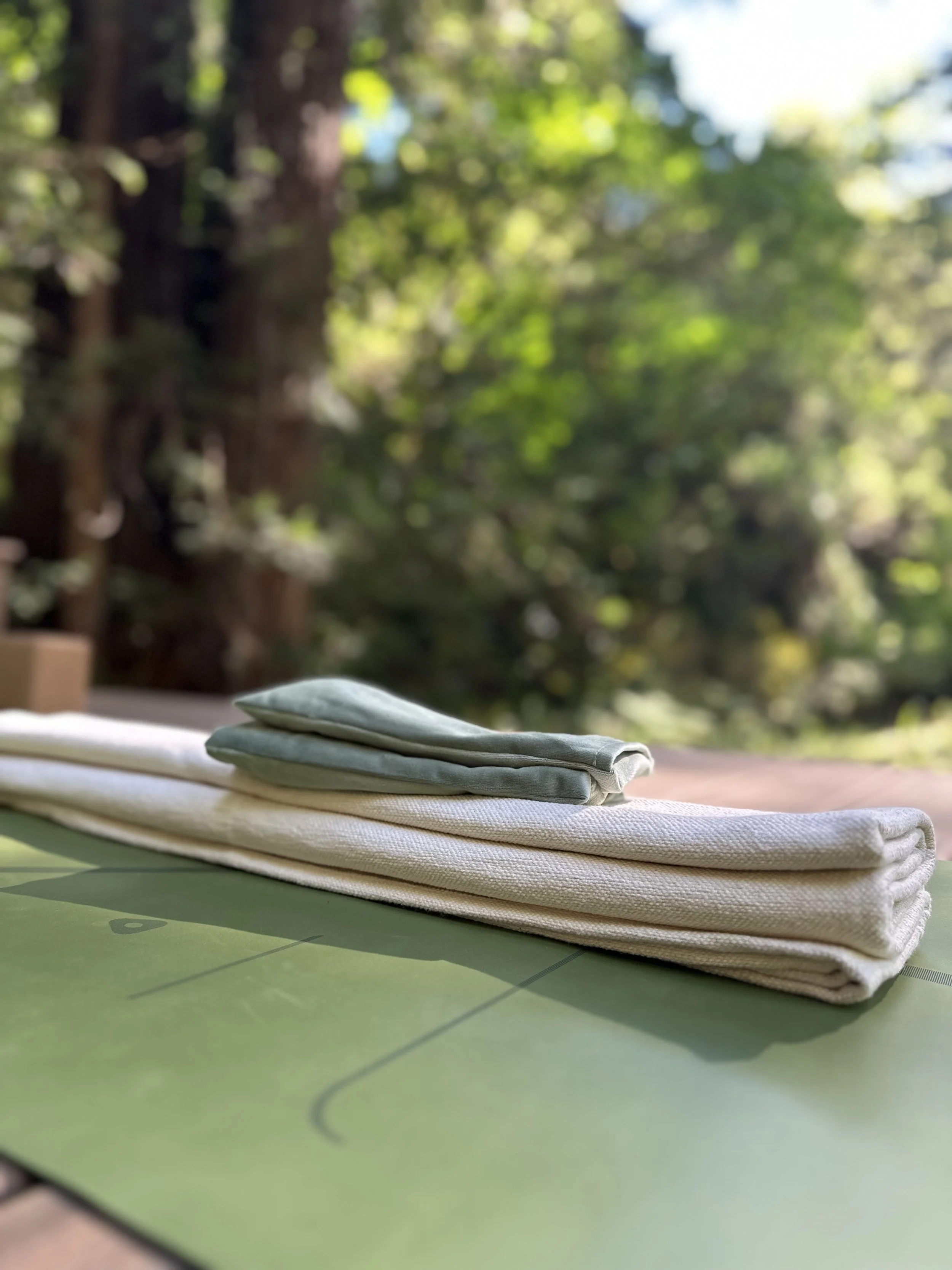 Folded towels placed on a green yoga mat outdoors with a blurred background of trees and sunlight.