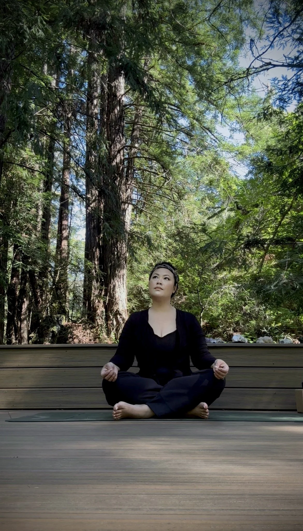 A woman practicing meditation outdoors on a wooden deck surrounded by tall trees and greenery, sitting cross-legged with her hands resting on her knees.