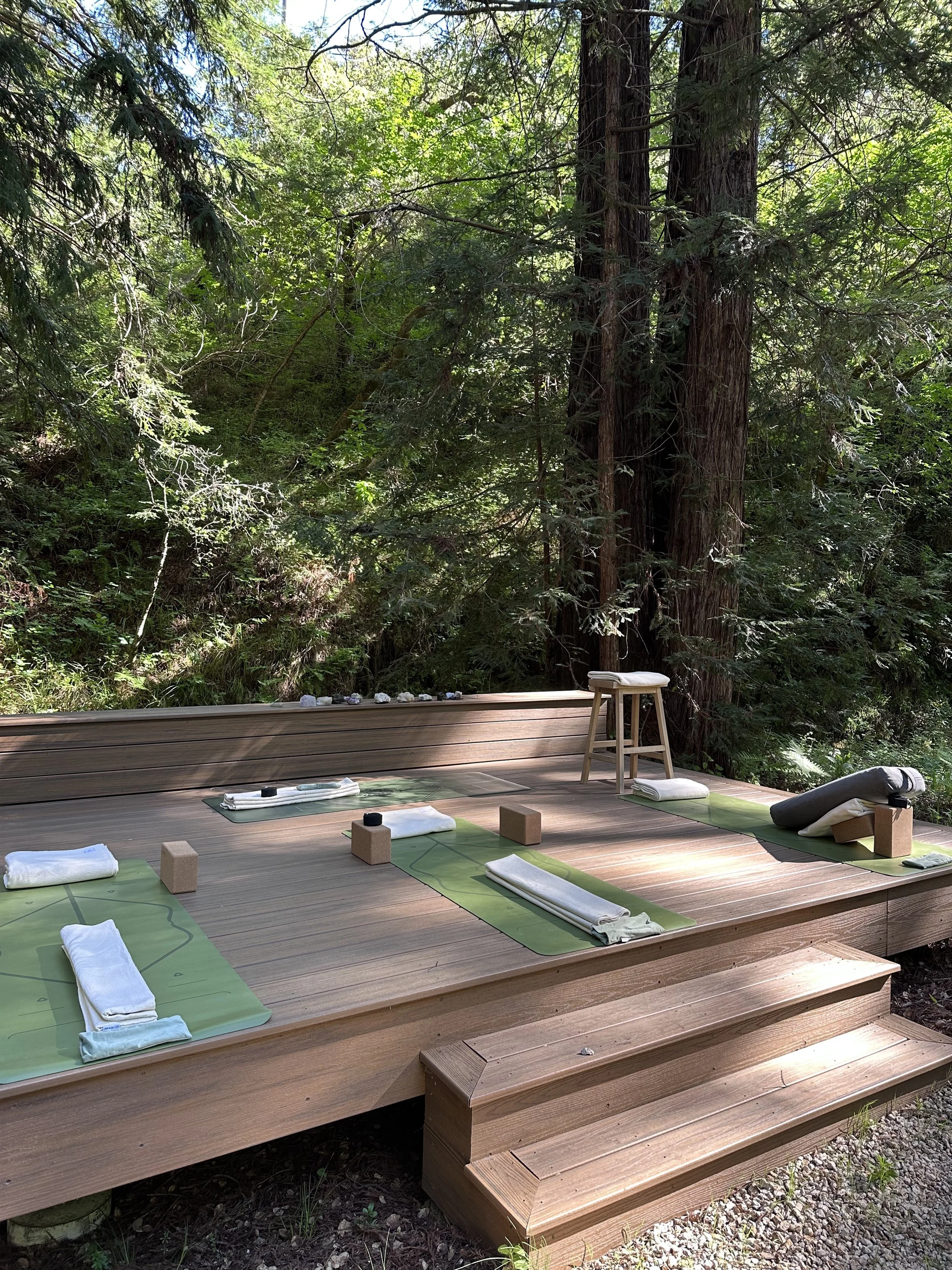 A wooden deck with yoga mats, towels, and blocks arranged for outdoor yoga practice, surrounded by trees and greenery.