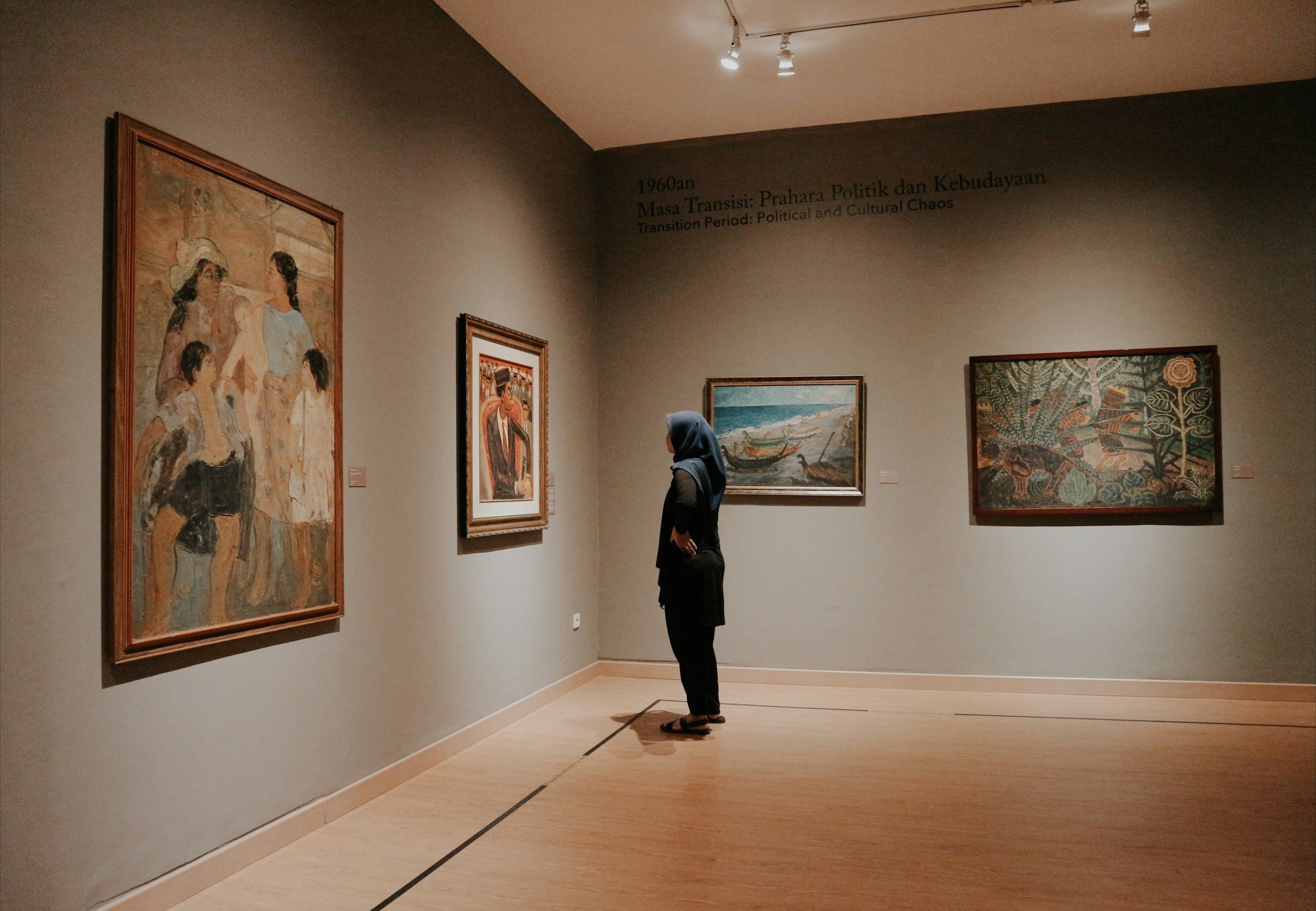 A woman wearing a blue hijab looking at paintings in an art gallery, with artworks depicting humans, landscapes, and traditional scenes.