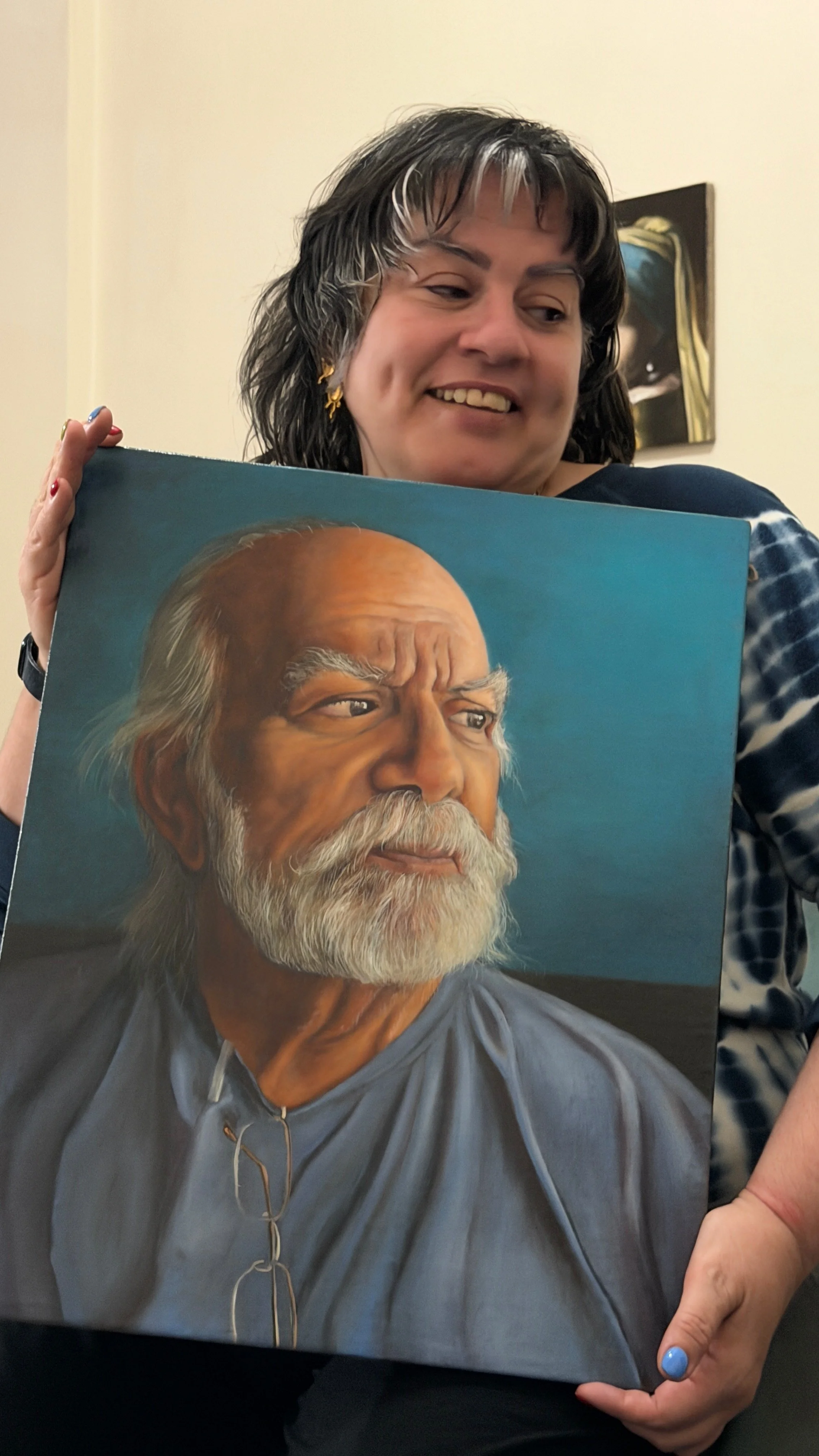 A woman holding a painting of an elderly man with white hair and a beard, wearing glasses and a gray shirt.