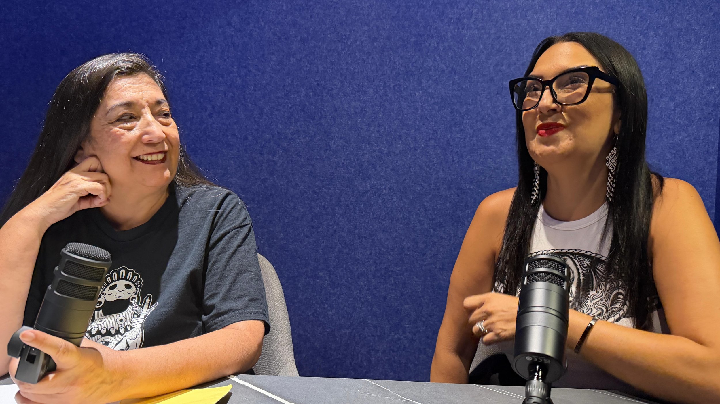 Two women sitting at a table with microphones, smiling and talking against a blue background.