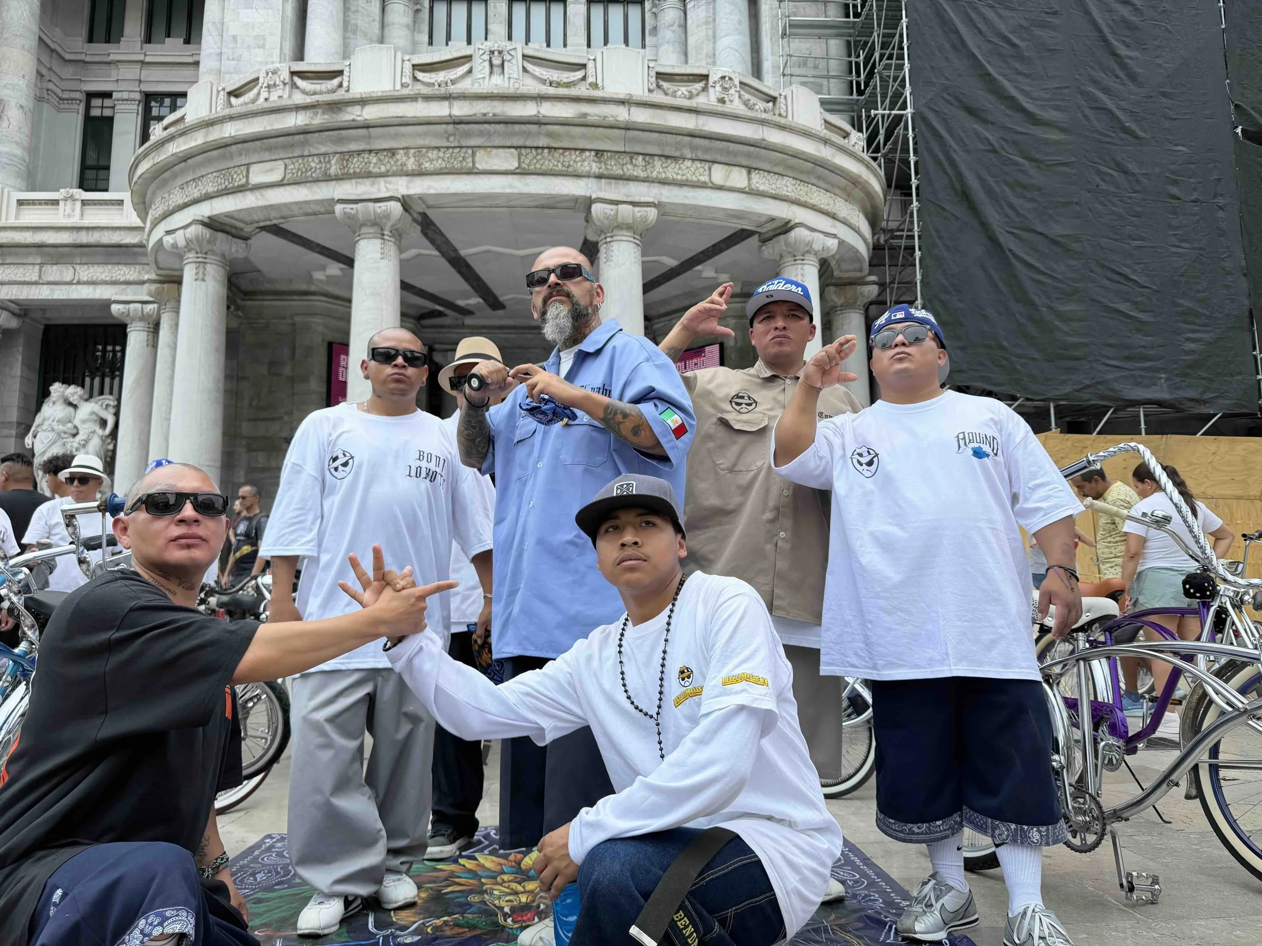 A group of people, some kneeling and some standing, gathered outdoors in front of a historic building with classical architecture. They are wearing casual clothing and sunglasses, with some making hand gestures. There is a black and gold helmet on the ground, and bicycles are visible. The scene appears to be at a public event or gathering.