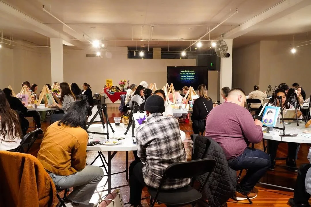 A group of people participating in a painting class in a spacious room with white walls and a wooden floor. Each person is seated at a table with painting supplies and easels, working on individual canvas paintings. There is a large TV screen at the front displaying text, and floral arrangements on a table at the back.