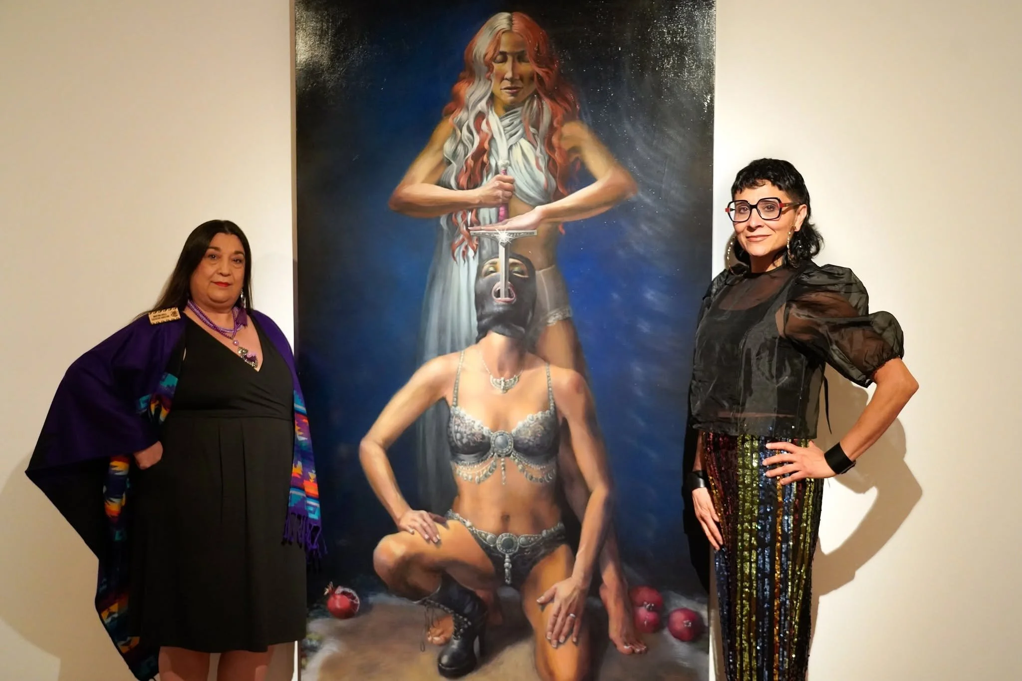 Two women stand beside a large fantasy-style painting of a woman with multicolored hair, armor, and a sword. The woman on the left wears a dark dress with a colorful shawl, and the woman on the right wears a black sheer top with patterned skirt. Both women pose beside the artwork in an art gallery.