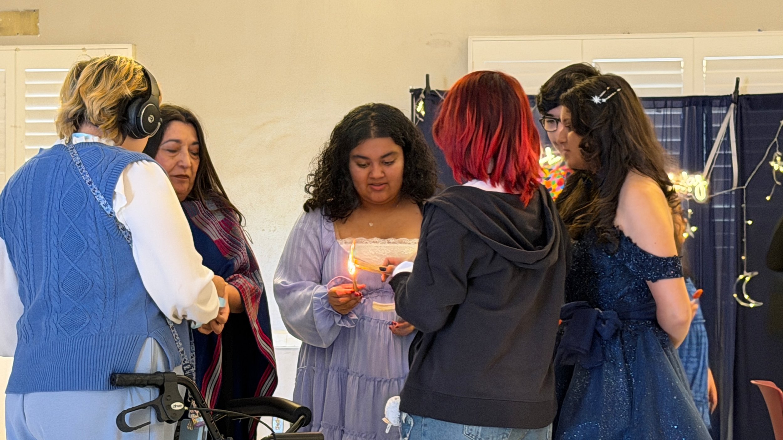 A group of five women gathered around a table holding a birthday cake with lit candles, in a decorated indoor space, with some women looking at the cake and others at each other.
