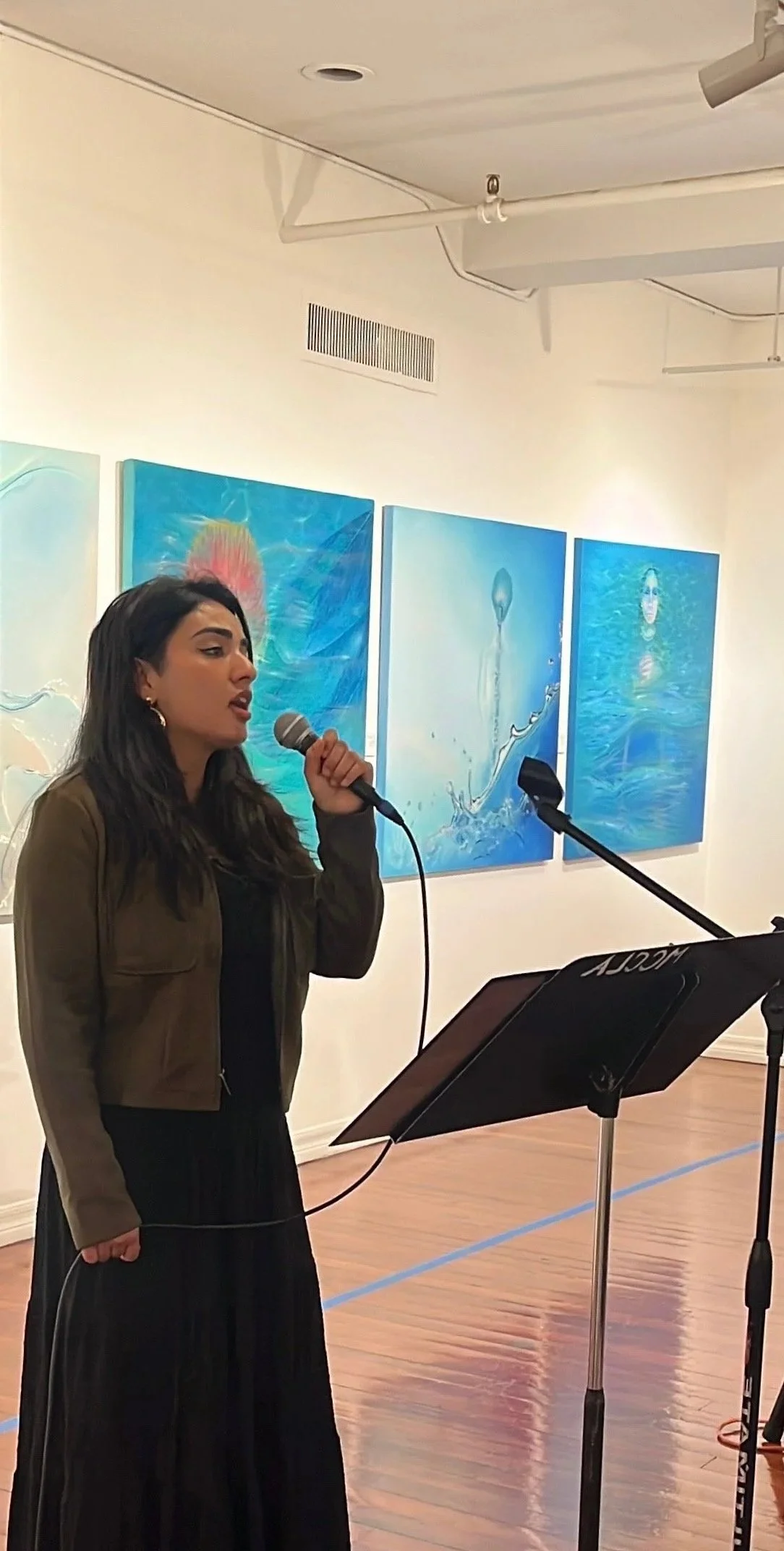 A woman singing into a microphone in an art gallery with three blue ocean-themed paintings in the background.