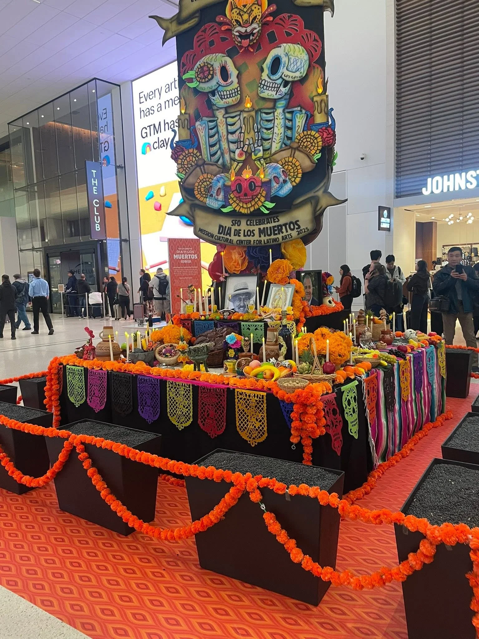 A colorful Día de los Muertos altar with marigold flowers, candles, framed photos, and decorative items, set up in an indoor space with people in the background.