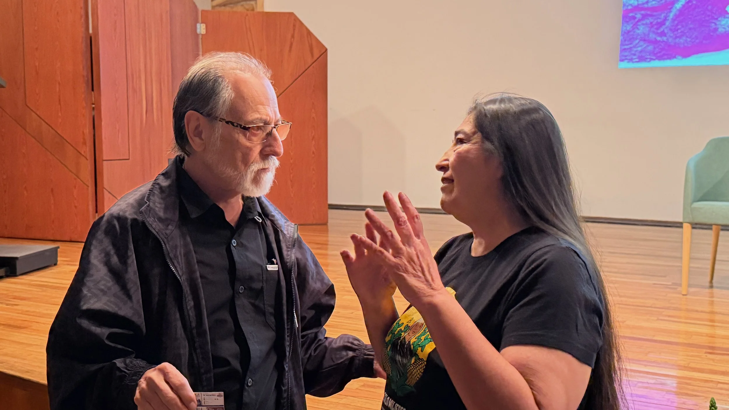 A man with white hair, glasses, and a beard wearing a black jacket and a woman with dark hair wearing a black shirt and colorful earrings are having a conversation, smiling, in an indoor setting with wooden floors and a colorful screen in the background.