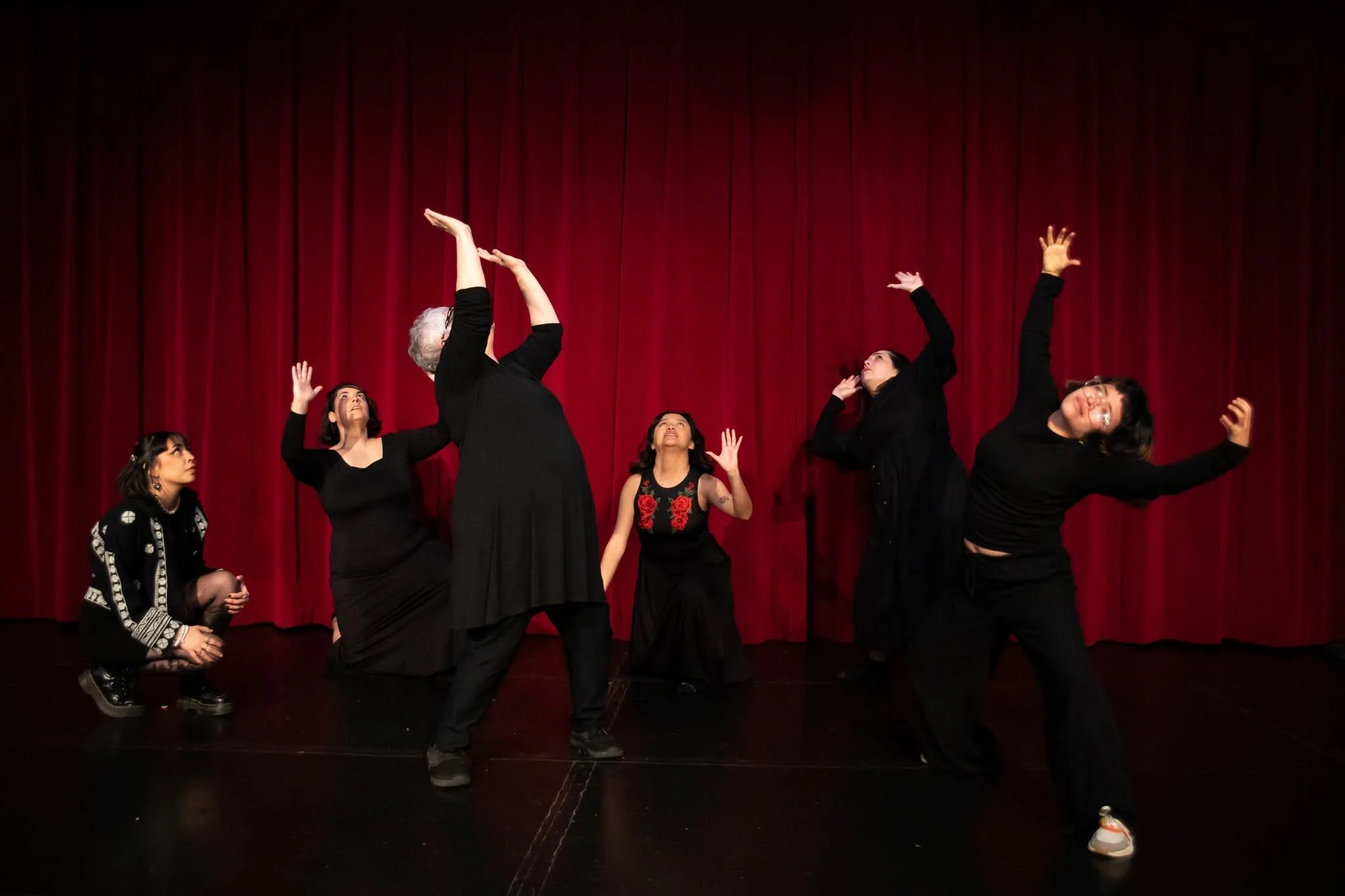 A group of six women performing a dance or theatrical act on stage in front of a red curtain.