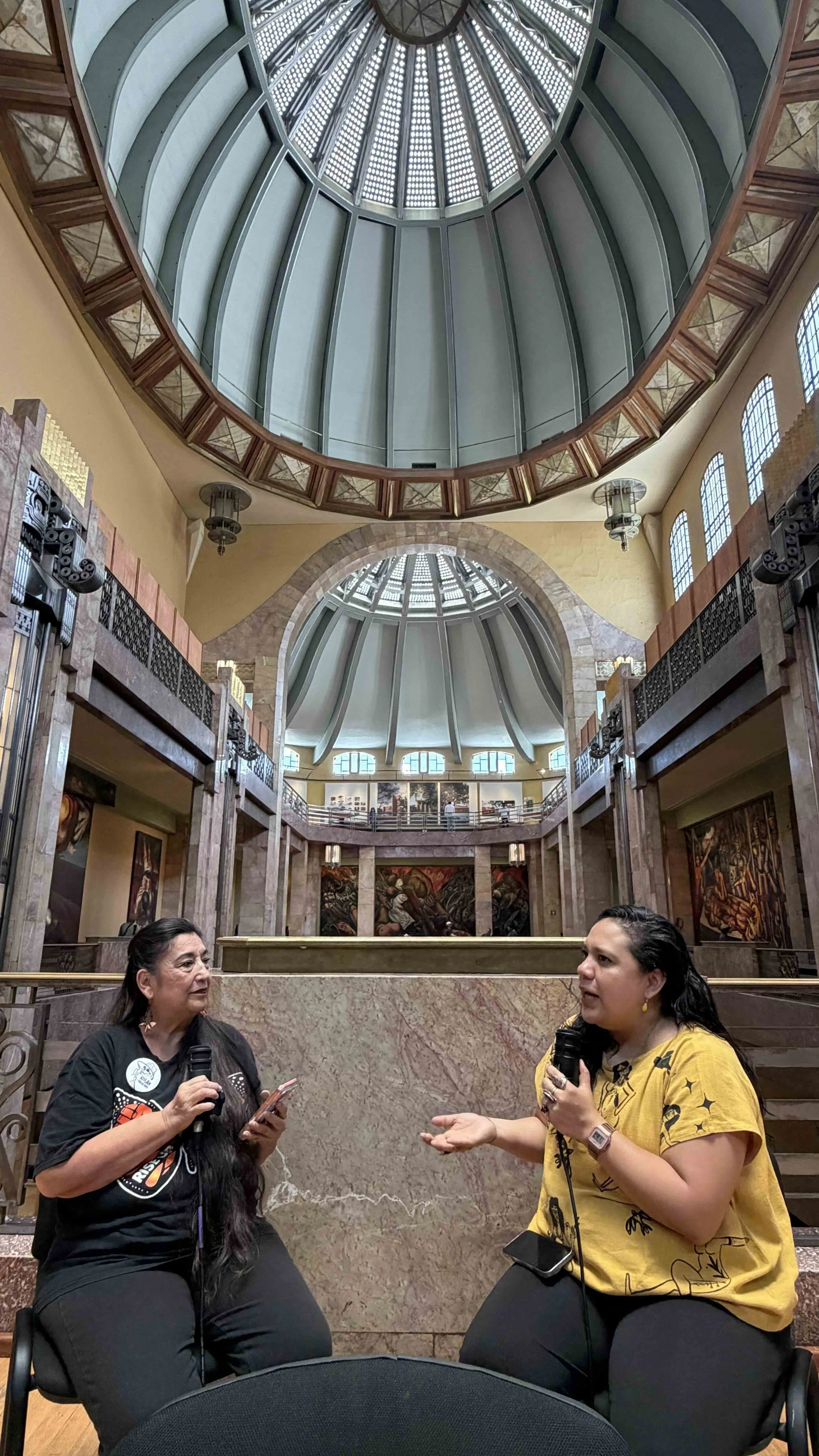 Two women sit in Palacio de Bellas Artes.  They are engaged in conversation, each holding microphones. One woman wears a black shirt with a graphic and the other a yellow shirt with black print.