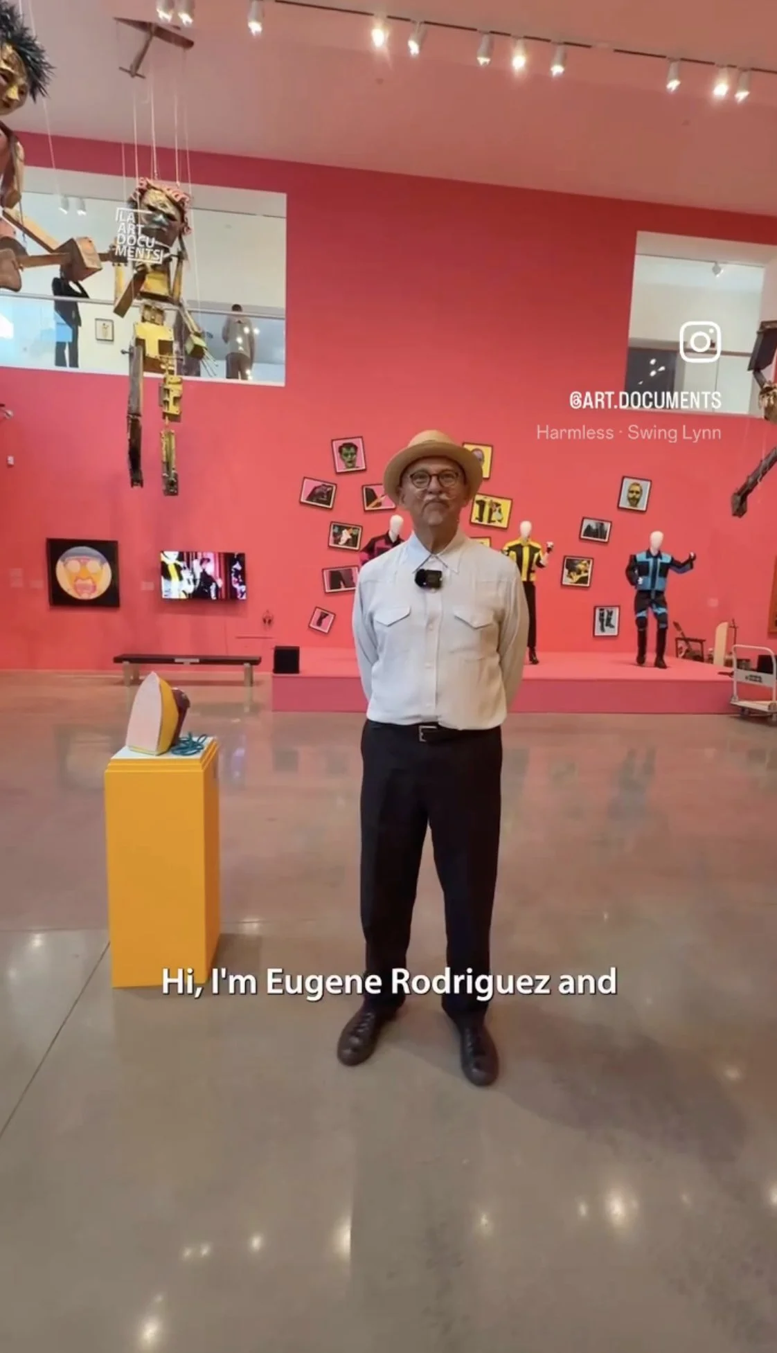 Eugene Rodriguez standing in an art gallery with pink walls, photo displays, mannequins, and art installations, introducing himself with subtitles.