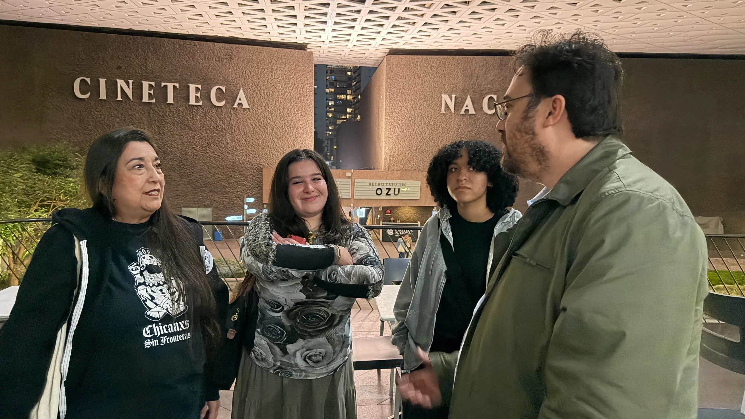 Four people standing and talking outside at night in front of a sign that reads 'Cineteca NAC.' The group includes two women and two men, one woman is wearing a black jacket with a graphic t-shirt, the woman next to her is smiling and has long dark hair, the young woman is wearing a gray hoodie, and the man has glasses and a green jacket. In the background, there are tall buildings with lights on, and a sign that says 'Retrosuizo Ozu.'