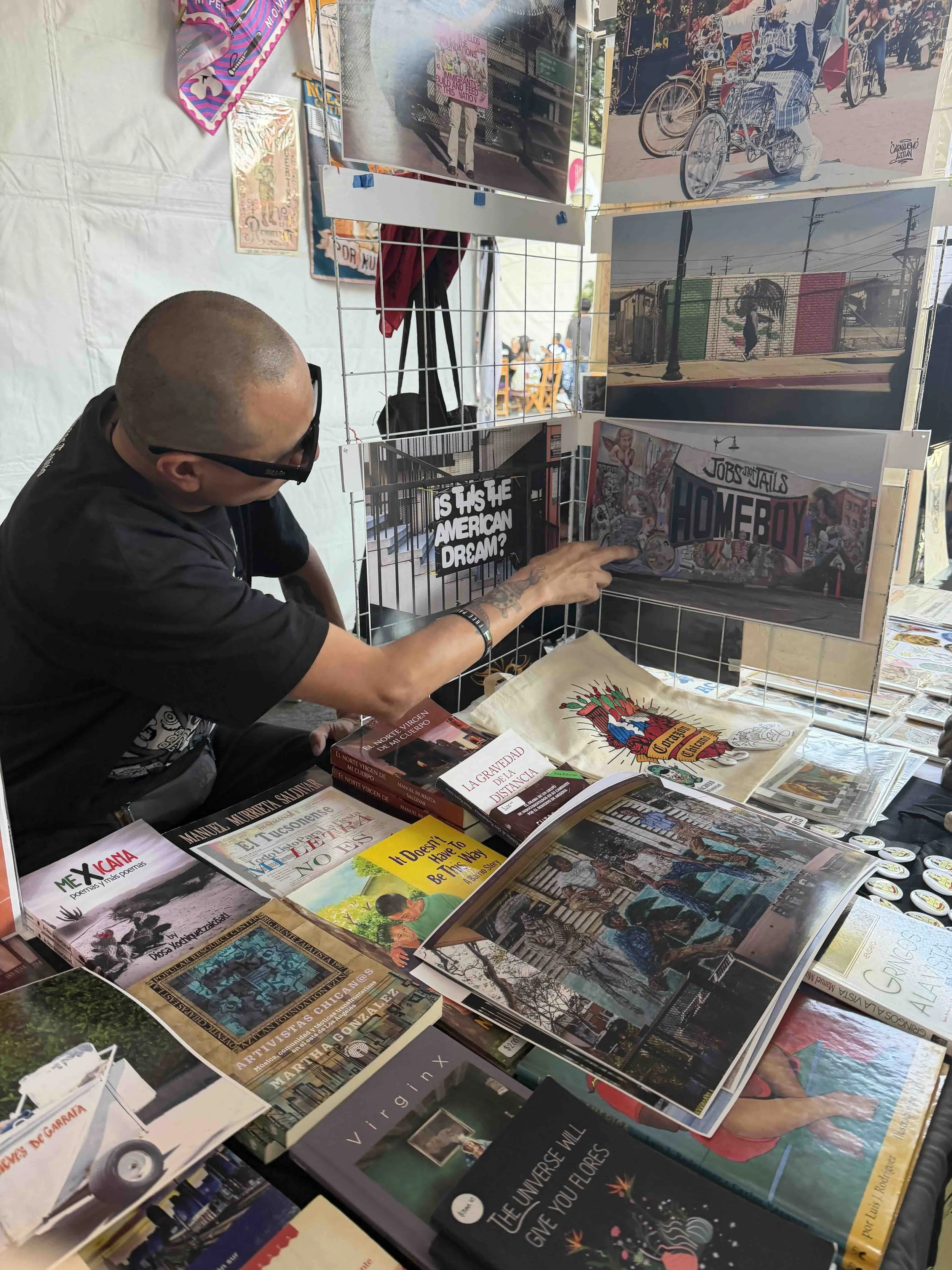 A person with a shaved head, wearing glasses, looking at books and art prints at a street market stall. The stall has various paintings, books, and printed materials displayed on a wire grid and tables.
