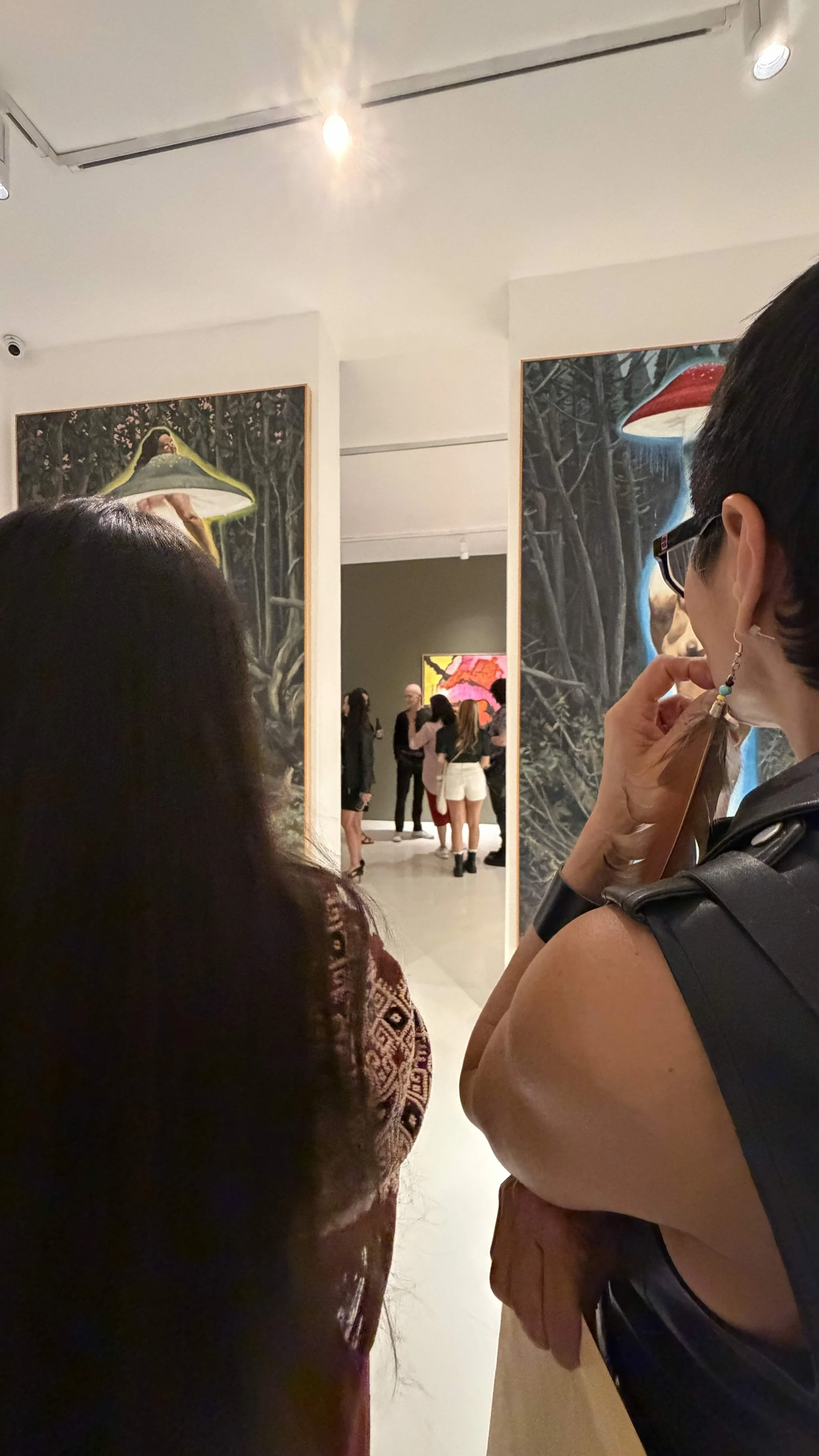 People viewing art in a gallery with large paintings of surreal forest scenes with mushrooms.