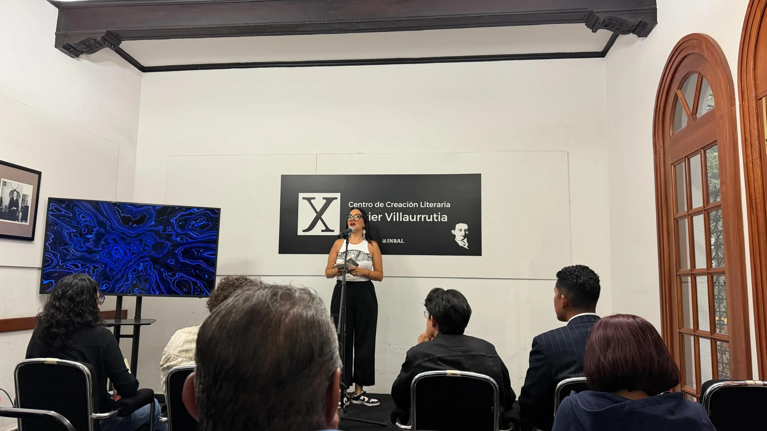 A woman wearing glasses and a sleeveless top holds a notebook and stands at a microphone in front of an audience in a room with a high ceiling, large windows, a black and white portrait, a large screen displaying a blue abstract pattern, and a black sign reading "Centro de Creación Literaria Xavier Villaurrutia".